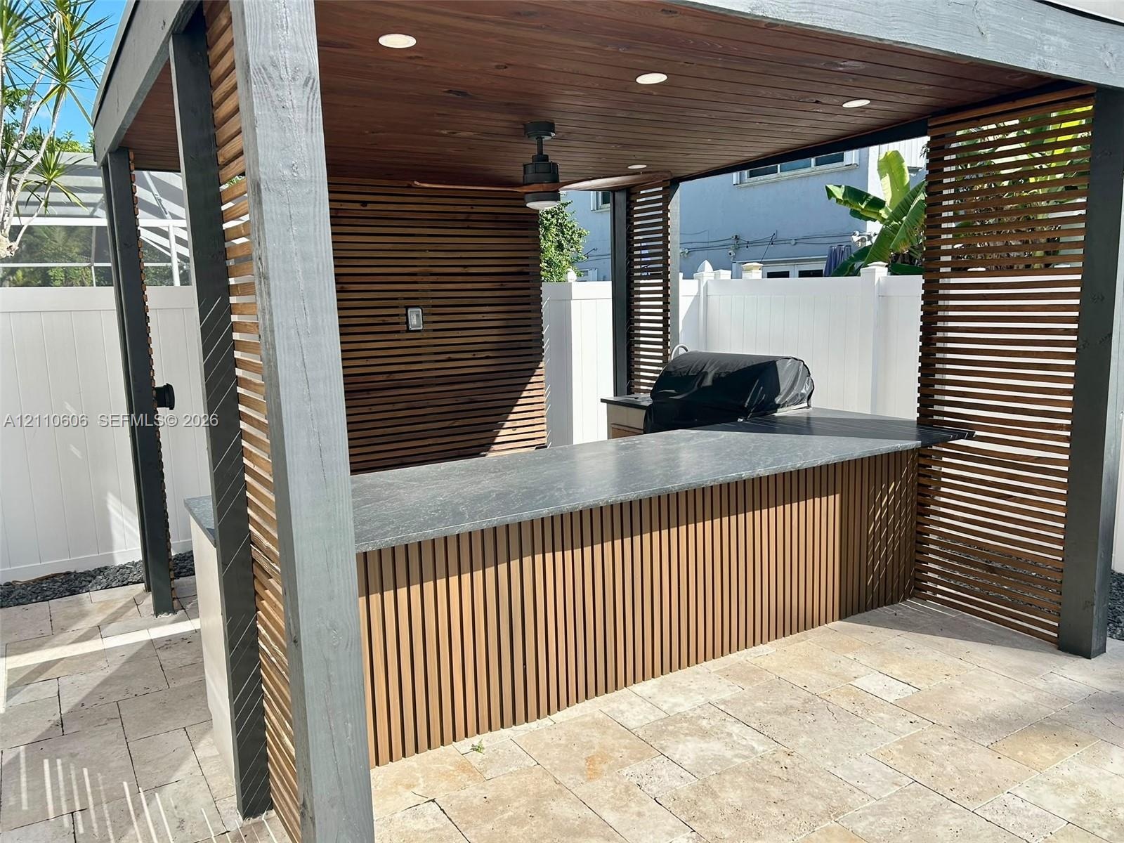 Covered outdoor kitchen featuring horizontal wood slat privacy walls, stone-finish bar top, recessed lighting, and a ceiling-mounted fan, situated on 