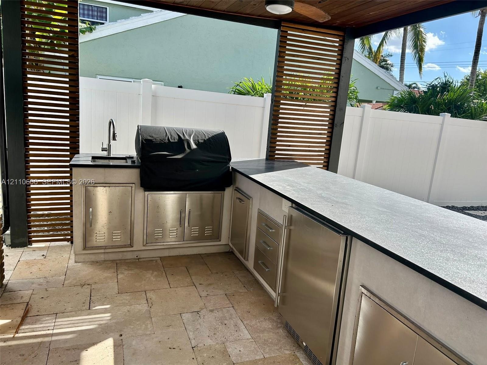 Covered outdoor kitchen featuring a stone-tile patio, built-in stainless steel cabinetry, a sink with a gooseneck faucet, and horizontal wood-slat pri