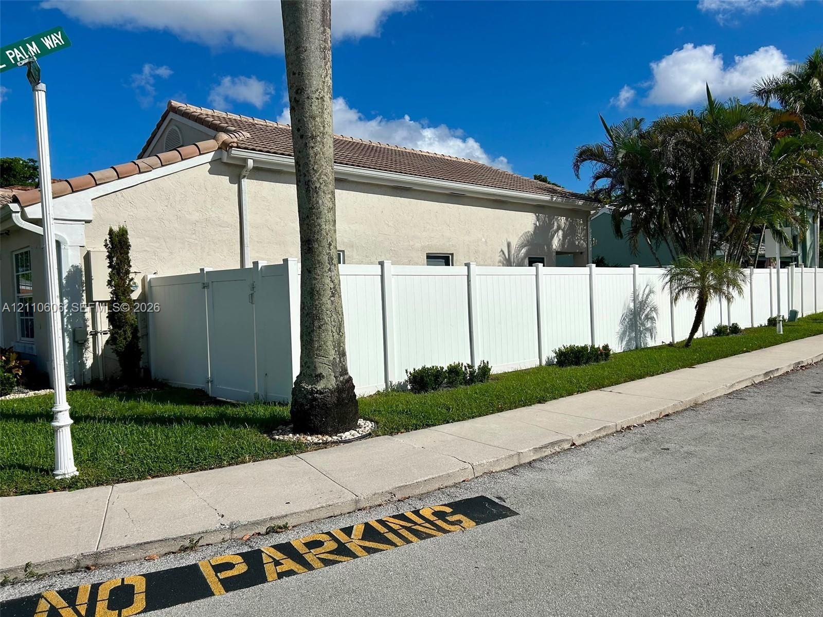 Exterior of property featuring a stucco facade, a tile roof, and a perimeter white privacy fence, surrounded by a landscaped lawn with palm trees