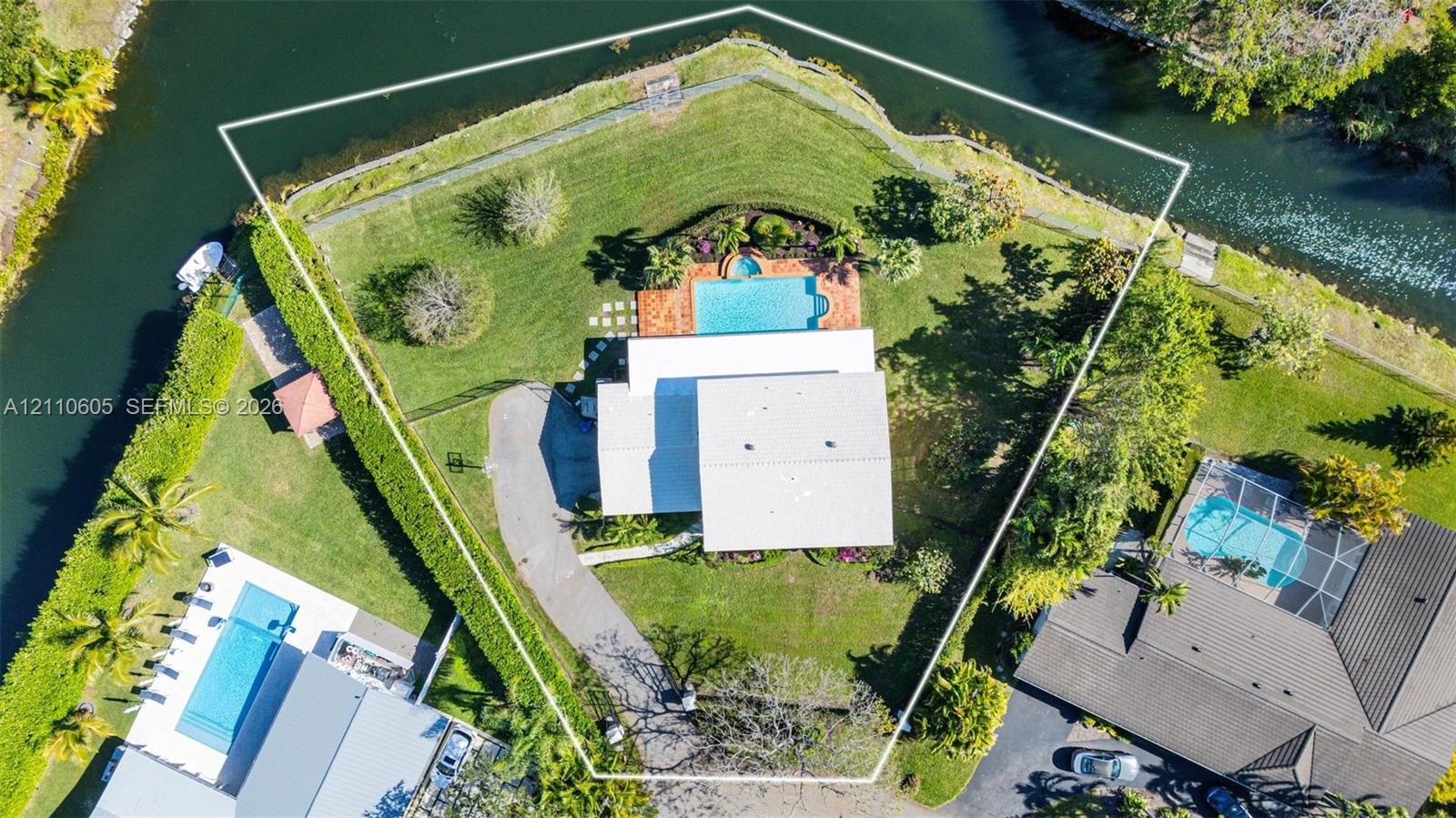 Waterfront lot featuring a perimeter fence, expansive lawn, mature landscaping, and a paved driveway leading to a swimming pool
