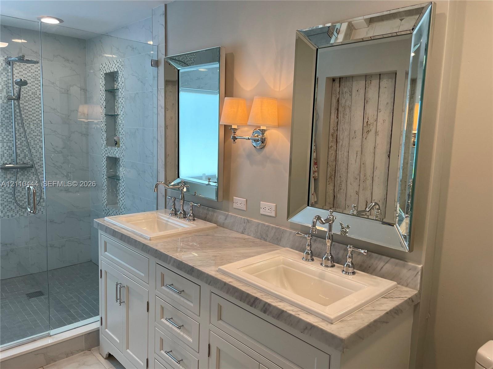 Double vanity featuring stone countertops, integrated rectangular sinks, polished metal widespread faucets, framed mirrors, and a walk-in shower with 