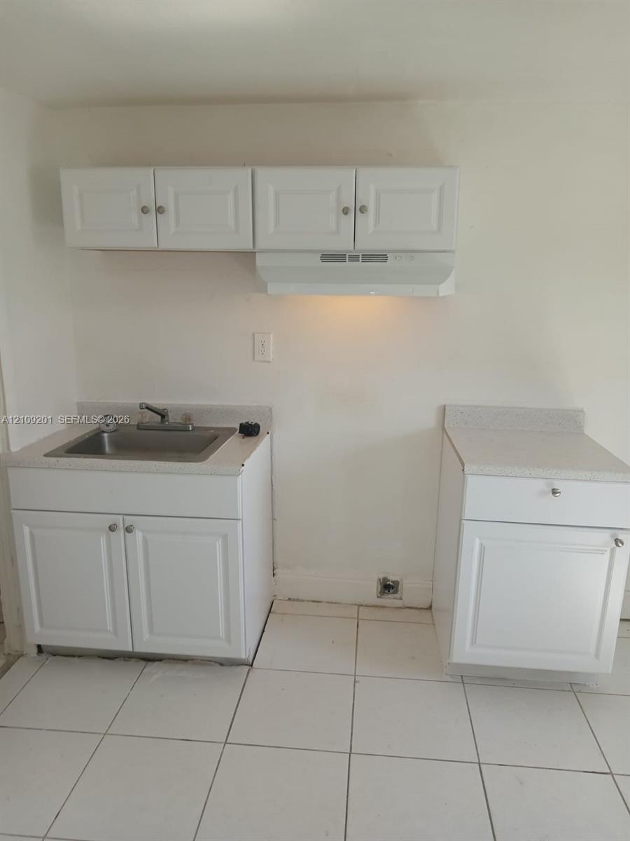 Kitchen area featuring white cabinetry, light-toned composite countertops, a stainless steel sink with a dual-handle faucet, a mounted range hood, and