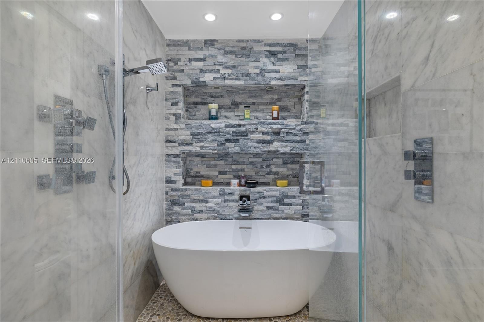 Freestanding soaking tub with polished chrome fixtures, integrated stacked-stone feature wall with dual recessed shelving, stone pebble flooring, and 