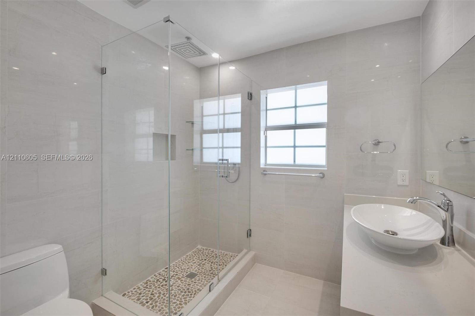 Bathroom featuring a glass-enclosed walk-in shower with pebble-stone flooring, a wall-mounted rainfall showerhead, a quartz-topped vanity with an oval