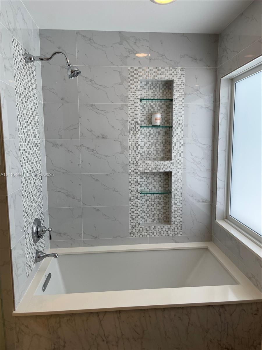 Bathtub surround featuring large-format veined tile, decorative mosaic tile accents, built-in recessed shelving with glass inserts, and polished chrom