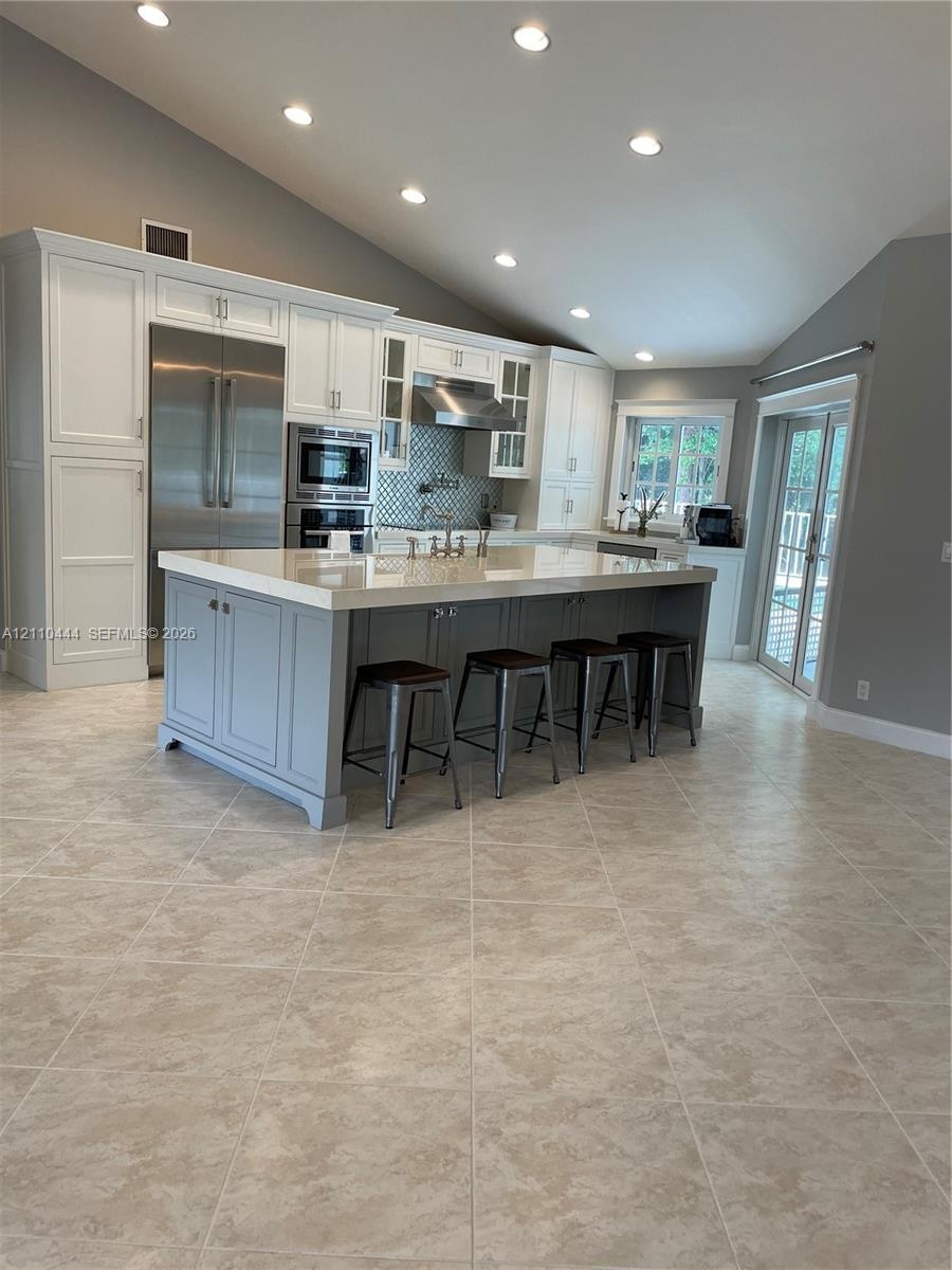 Kitchen featuring an oversized island with quartz countertops, custom cabinetry, stainless steel appliances, tile flooring, and vaulted ceilings with 
