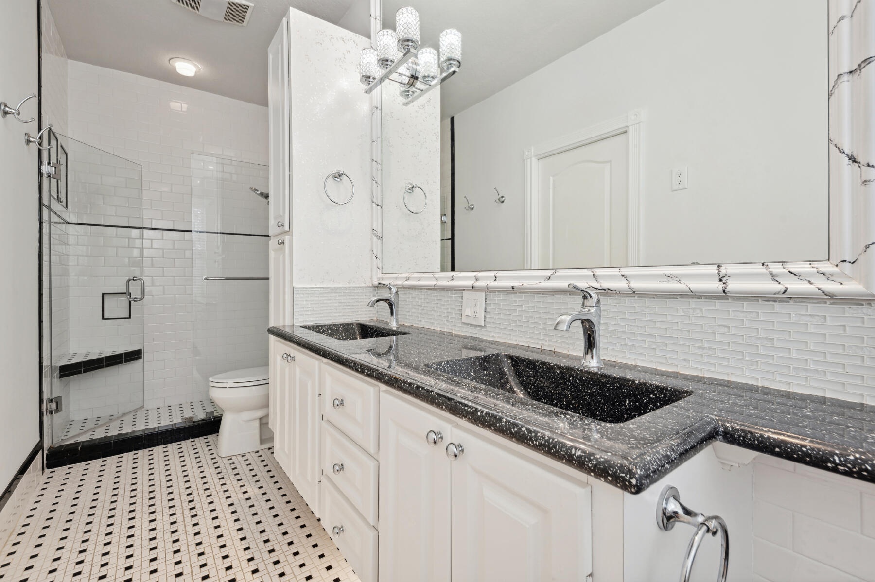 Double vanity with dark speckled countertop and integrated sinks, white cabinetry, glass-enclosed shower with subway tile surround, and mosaic tile fl