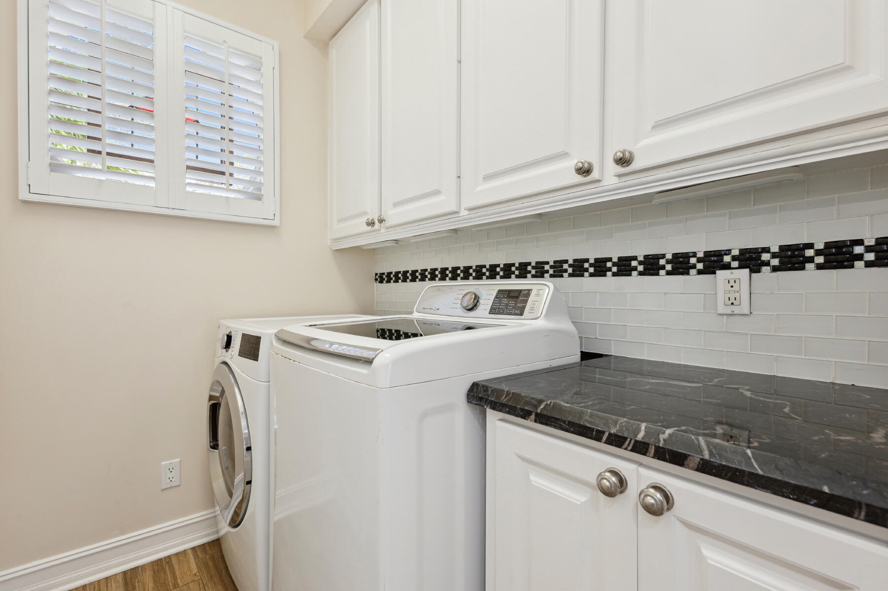 Laundry area featuring white shaker-style cabinetry, dark stone-finish countertops, subway tile backsplash with decorative inset, and plantation shutt