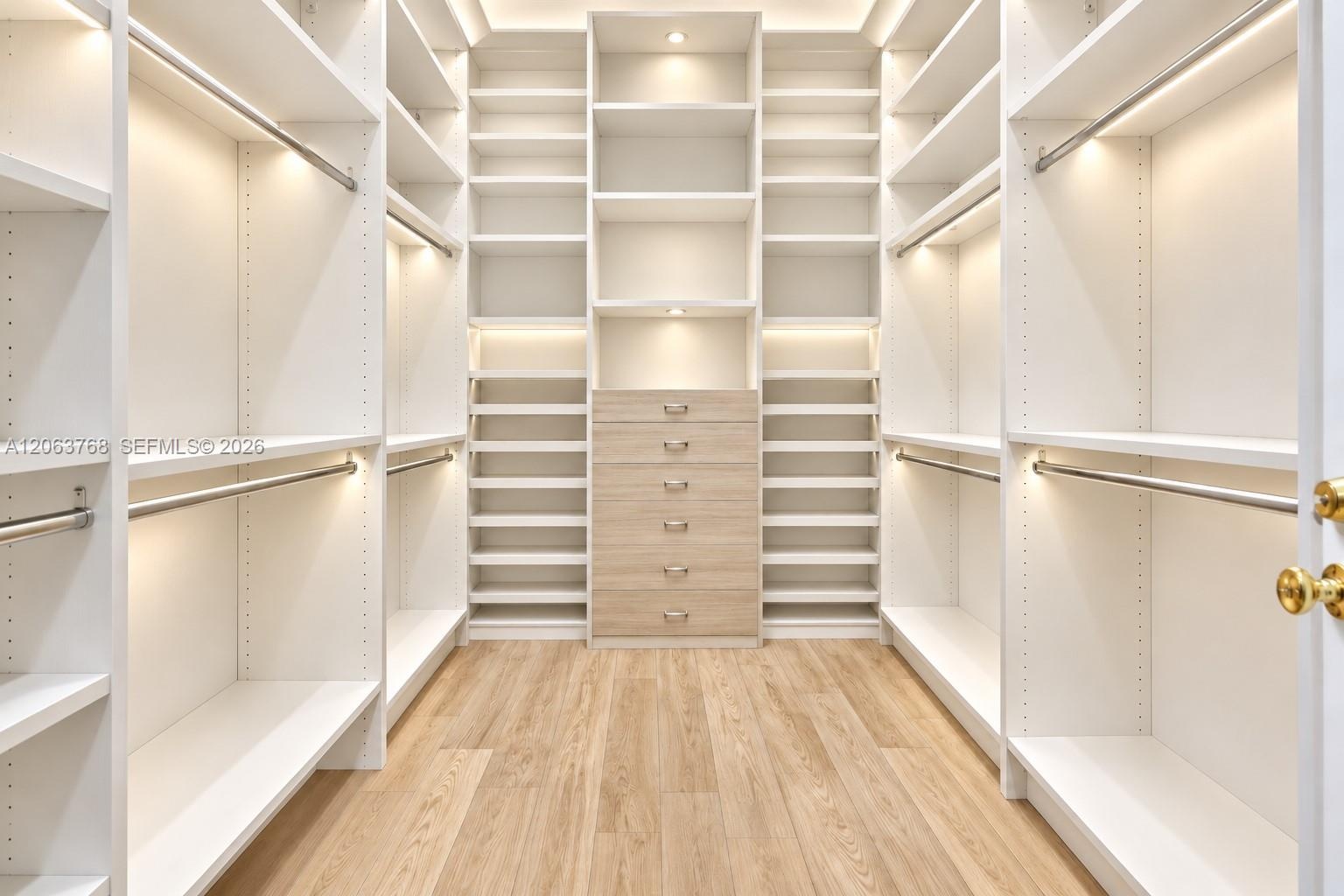 Custom walk-in closet featuring floor-to-ceiling white shelving, integrated recessed lighting, wood-finish flooring, and a central built-in chest of d