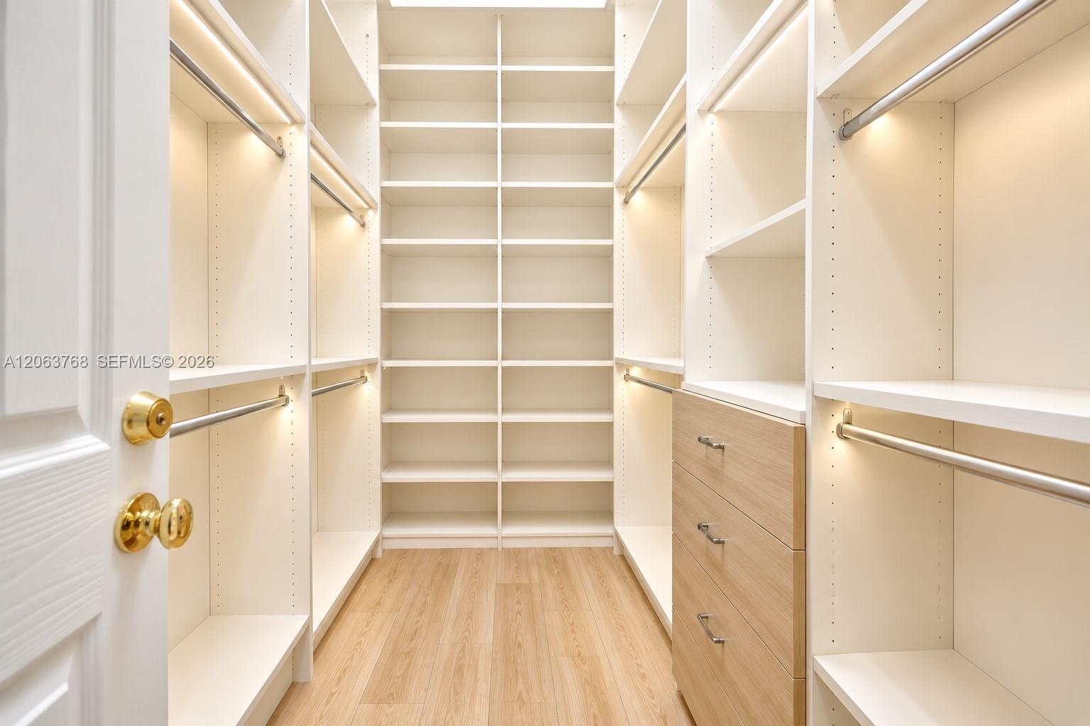 Walk-in closet featuring extensive built-in white shelving, integrated LED lighting, wood-finish drawer units with brushed metal hardware, and wood-fi
