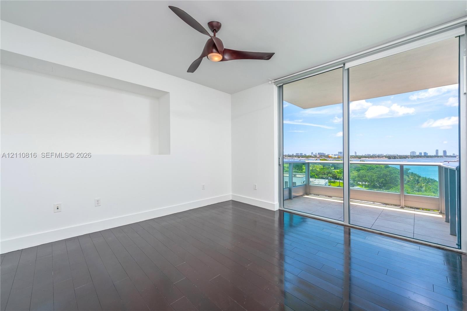 Dark wood-finish flooring, recessed wall niche, modern blade-style ceiling fan, and floor-to-ceiling sliding glass doors opening to a private balcony 