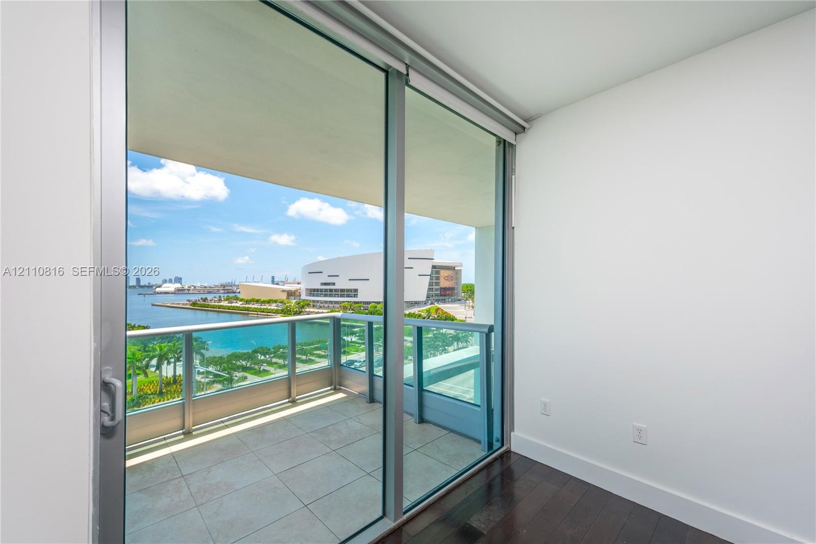 Floor-to-ceiling sliding glass door access to a covered balcony with a metal and glass railing, featuring tile flooring and hardwood-finish interior f