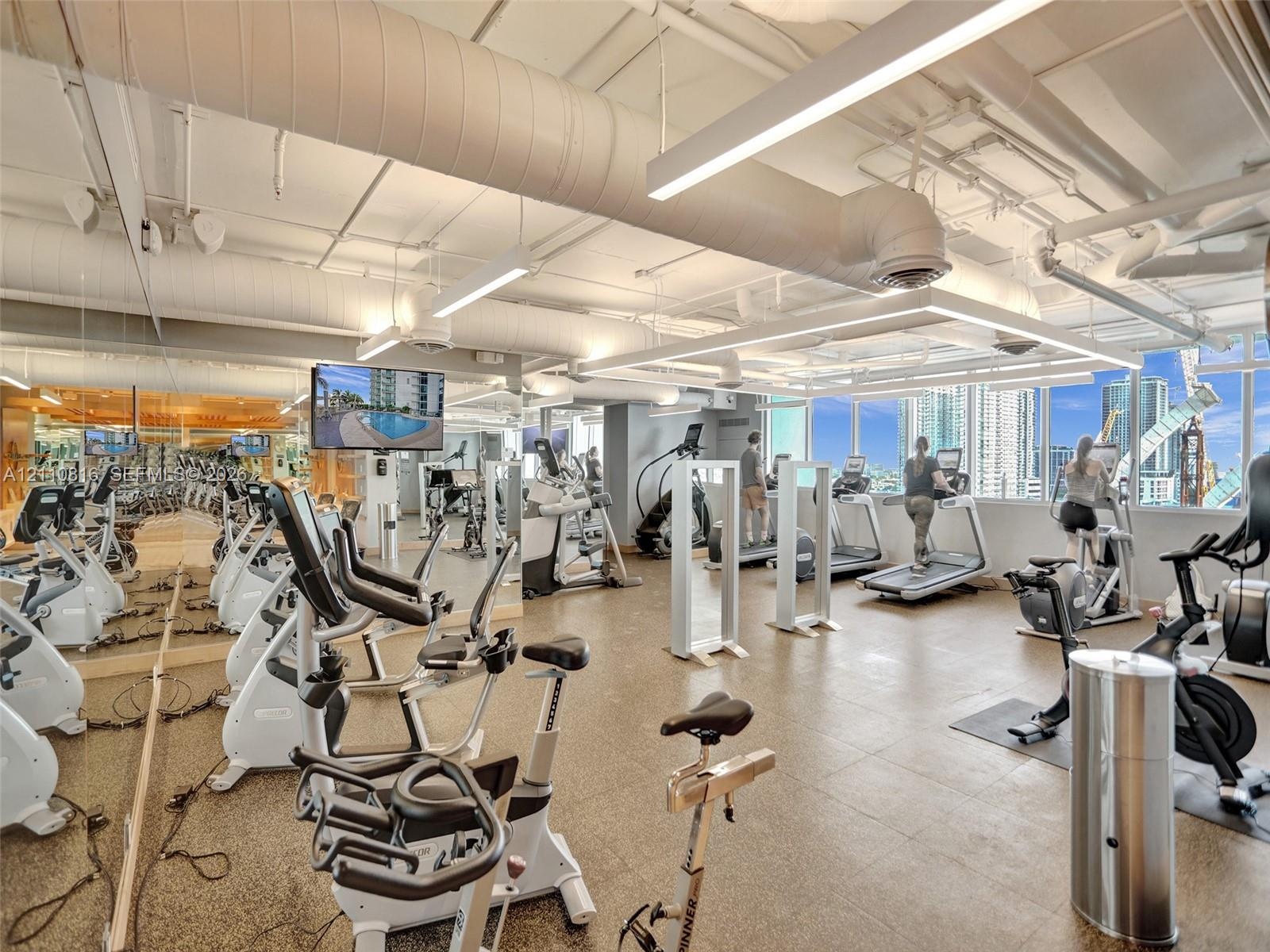 Fitness center featuring industrial-style exposed ductwork, wall-to-wall mirrored surfaces, expansive windows, linear overhead lighting, and stone-fin