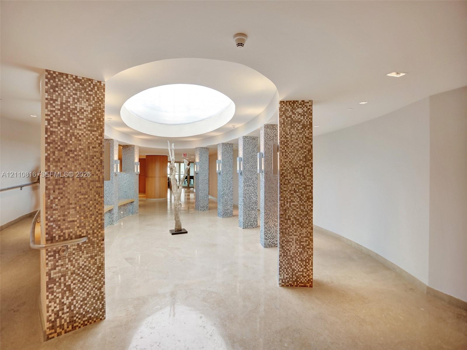 Circular skylight featuring a recessed domed ceiling, stone flooring, and structural columns finished with multi-toned glass mosaic tiling