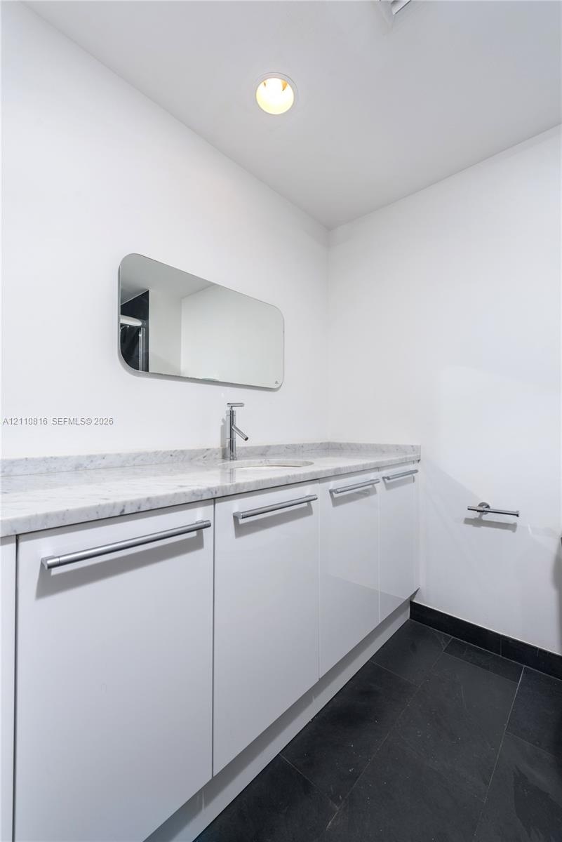 Custom white cabinetry with sleek horizontal pulls, stone-composite countertop, undermount sink, wall-mounted mirror with rounded edges, and dark tile