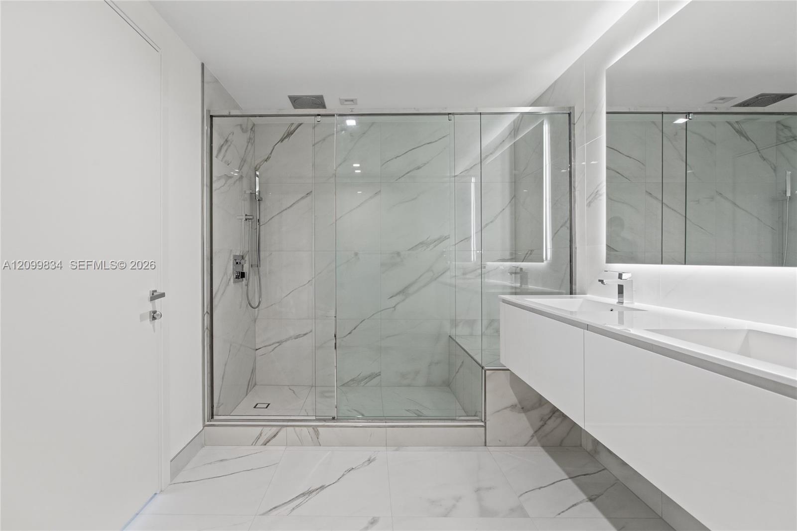 Bathroom featuring floor-to-ceiling veined stone-finish tile, glass-enclosed shower with built-in bench, floating dual-sink vanity, and expansive wall