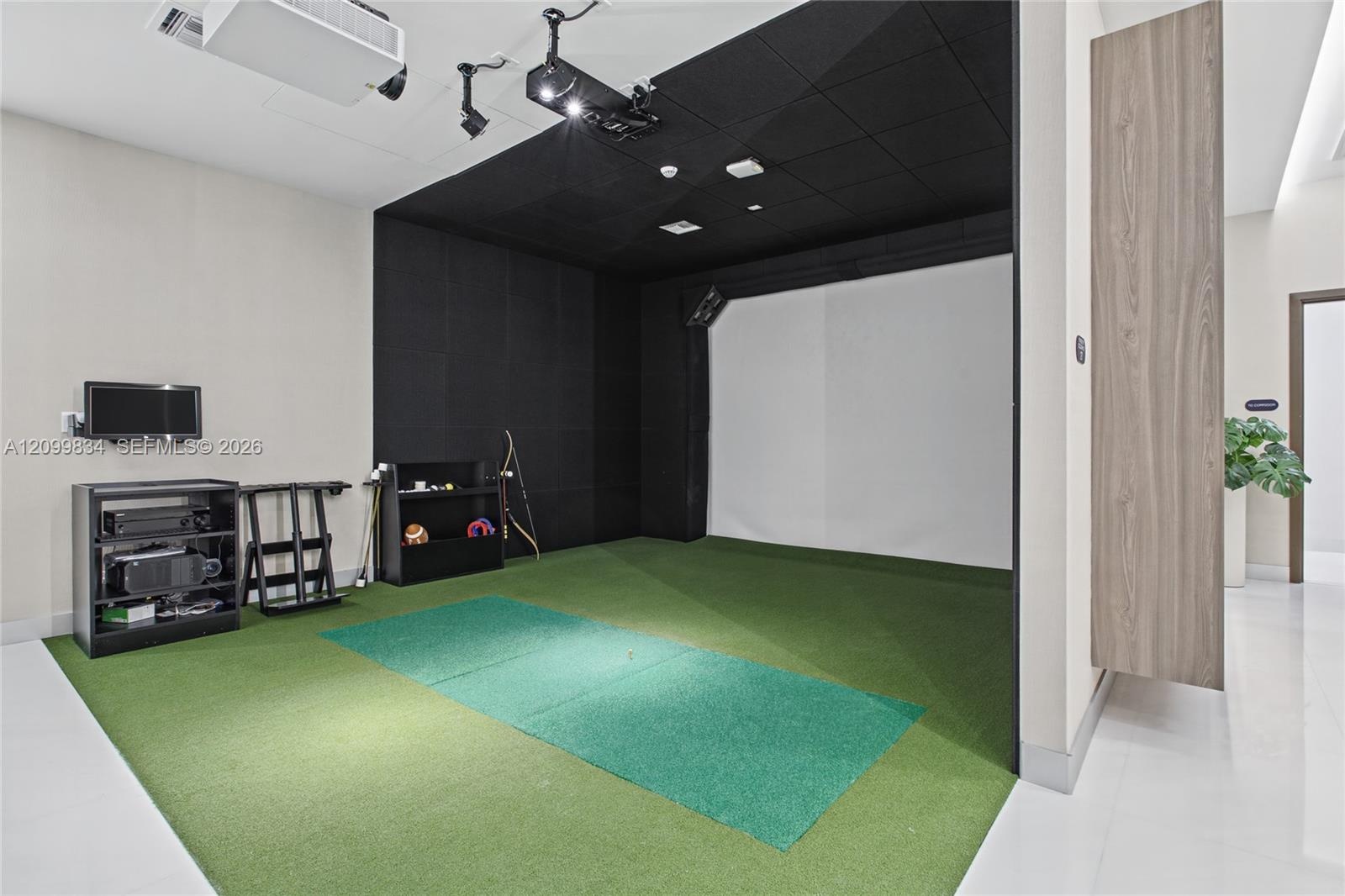Dedicated simulation room featuring synthetic turf flooring, floor-to-ceiling acoustic wall panels, an integrated projection screen, and overhead trac
