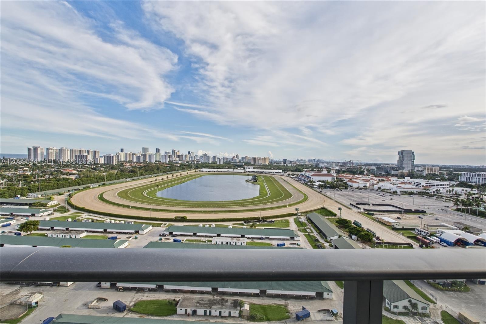 Elevated balcony with a dark metal railing overlooking a racetrack, central water feature, and urban skyline