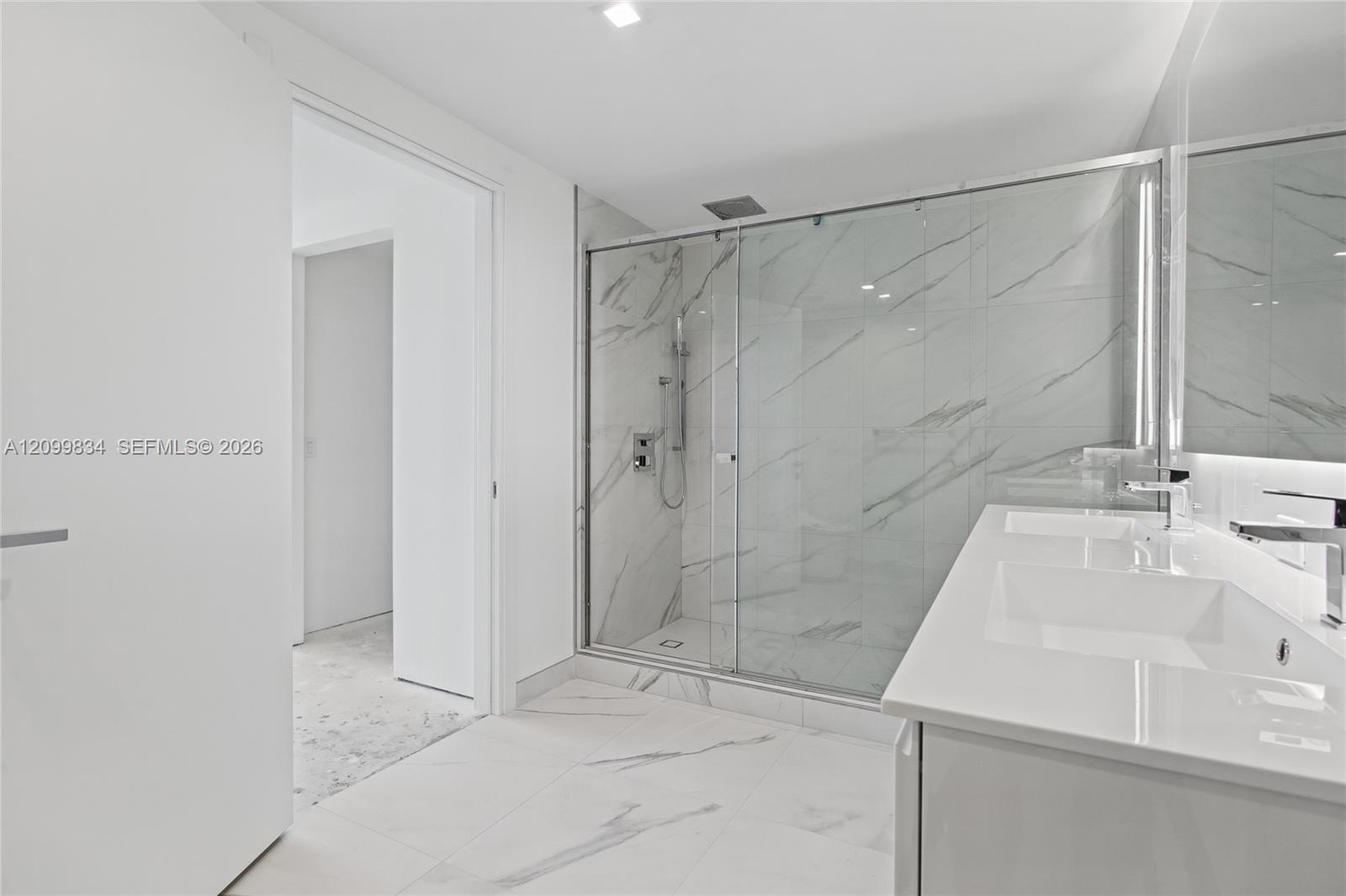 Bathroom featuring a dual-sink vanity with a white solid-surface countertop, walk-in shower with floor-to-ceiling stone-finish tile, glass shower encl