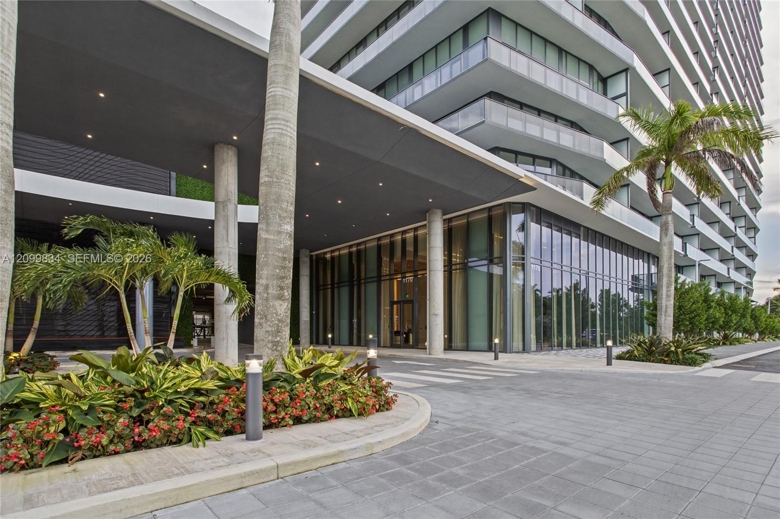Covered porte-cochere entrance featuring floor-to-ceiling glass paneling, concrete structural columns, recessed exterior lighting, and manicured tropi