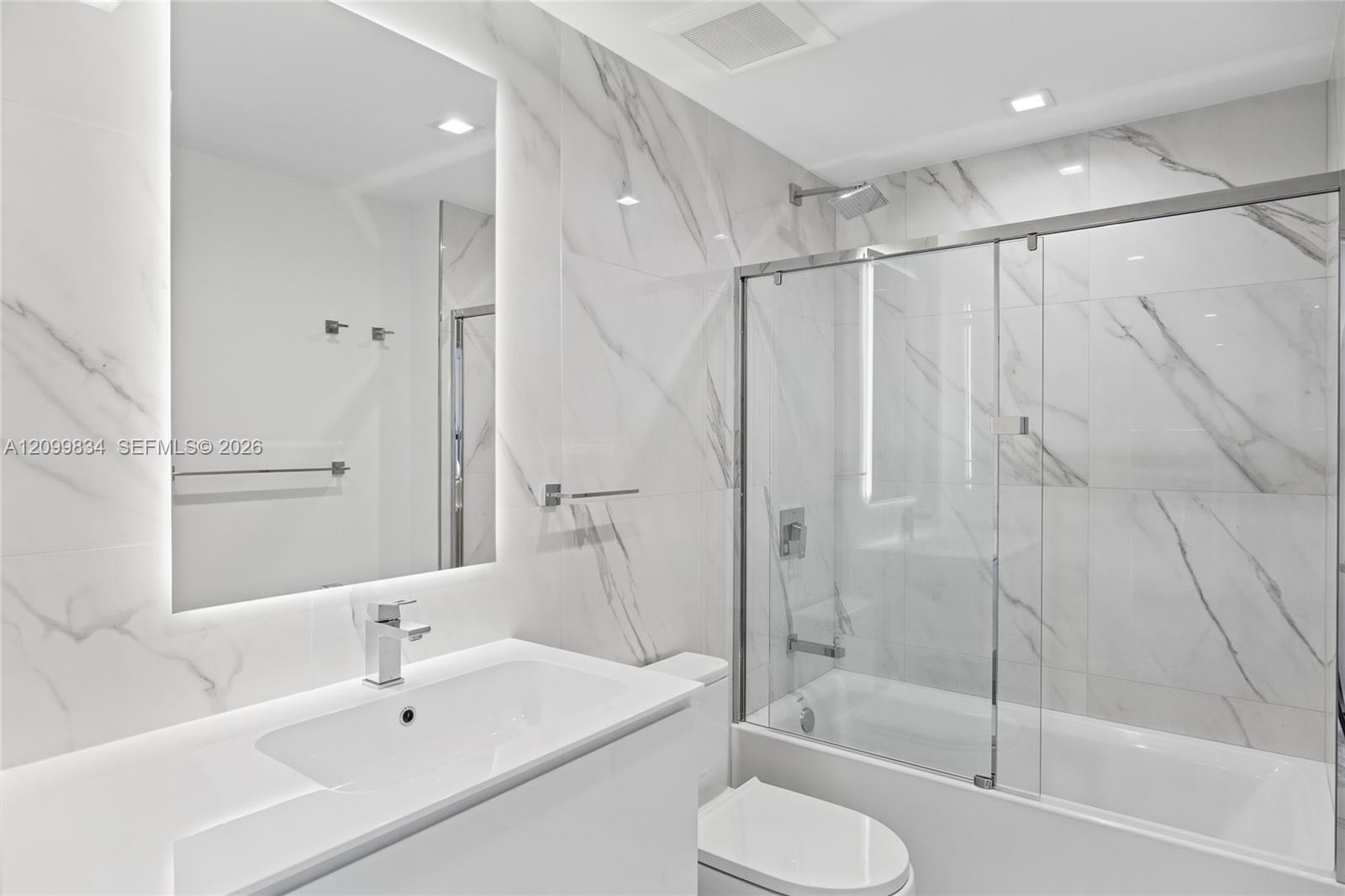 Bathroom featuring floor-to-ceiling large-format stone-finish tile, a backlit mirror, a vanity with a rectangular basin, and a glass-enclosed shower-t