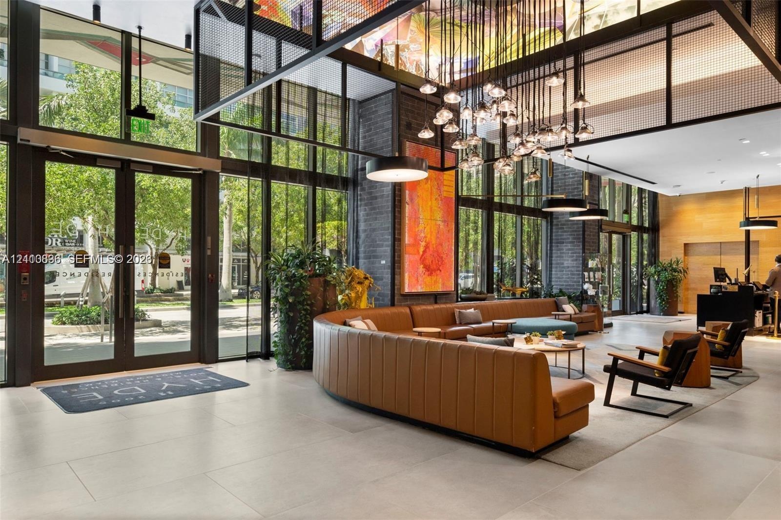 Double-height lobby featuring floor-to-ceiling glass walls, large-format tile flooring, exposed brick structural columns, and a custom multi-light cha