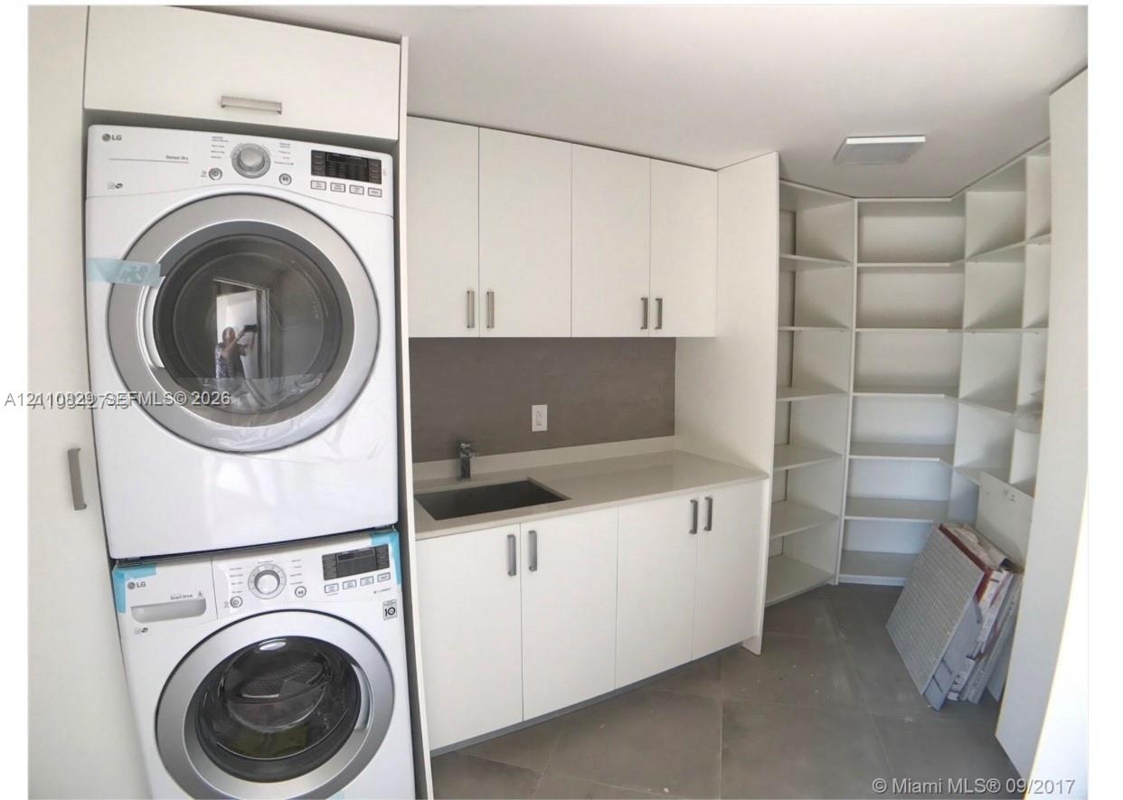 Utility area featuring white flat-panel cabinetry, a solid-surface countertop, an integrated sink with a chrome faucet, a stacked washer and dryer uni