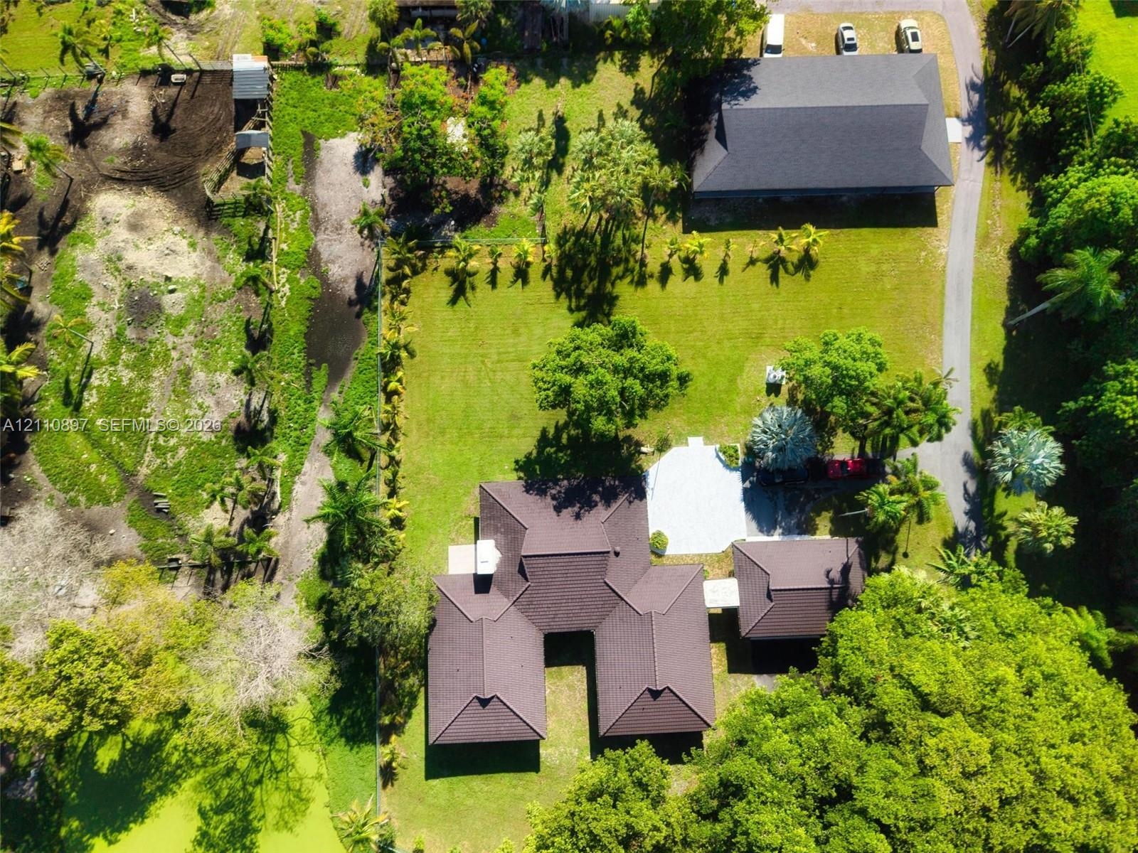 Aerial perspective featuring a sprawling lot with mature trees, a detached multi-vehicle structure, a paved driveway, and a residence with a multi-hip