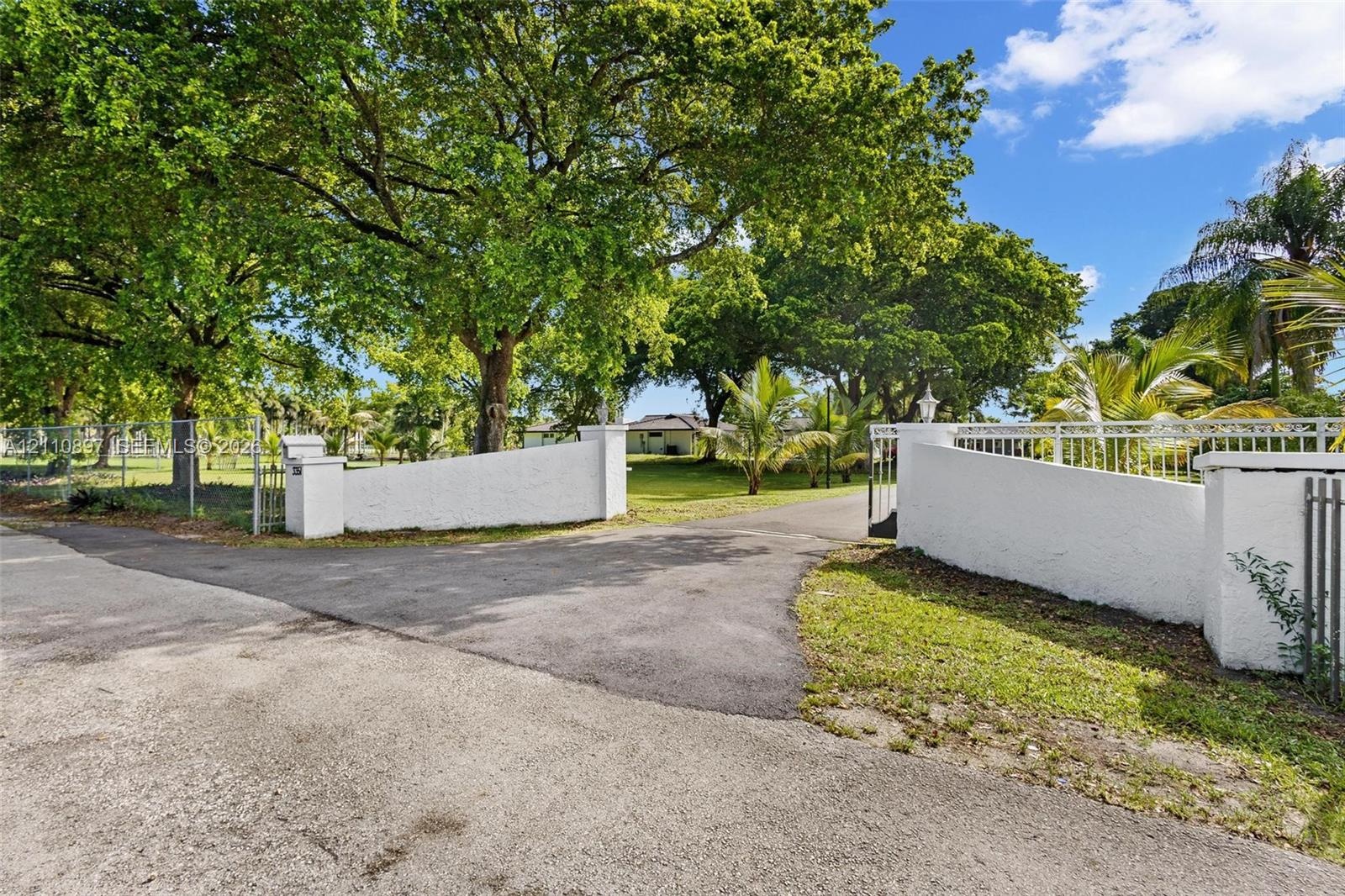 Gated entry featuring stucco-finished masonry pillars, a paved driveway, and mature canopy trees