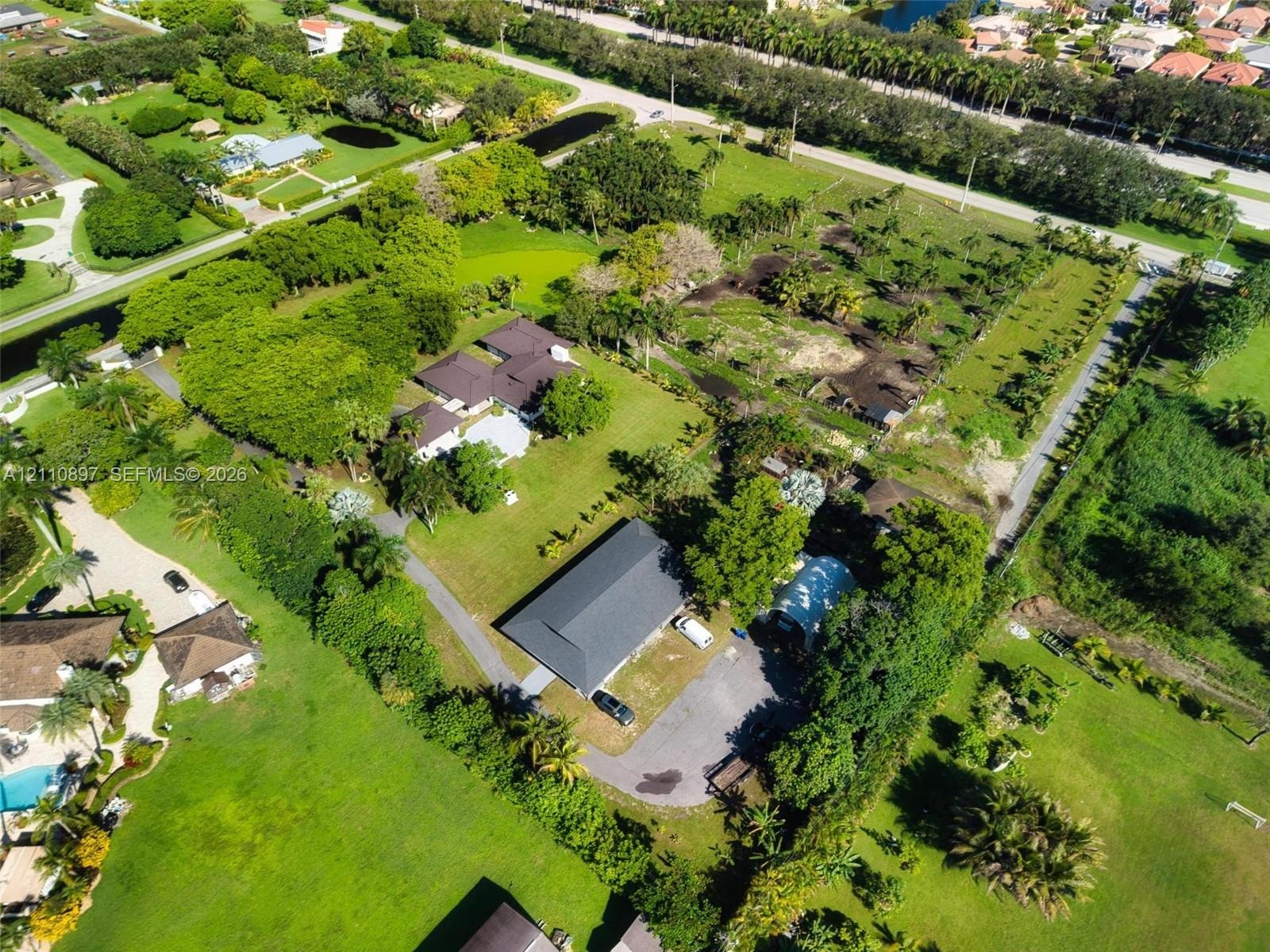 Aerial perspective of a sprawling property featuring extensive open green space, mature canopy trees, asphalt driveways, and multiple residential stru