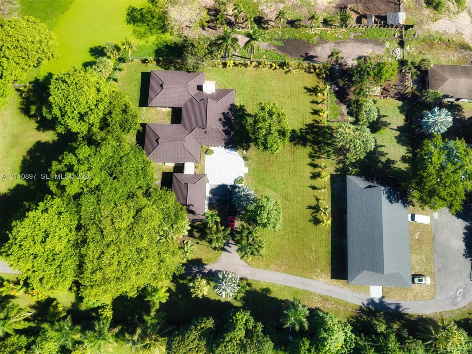 Aerial perspective of a sprawling property featuring mature canopy trees, a dedicated secondary structure with a dark shingle roof, paved driveway, an