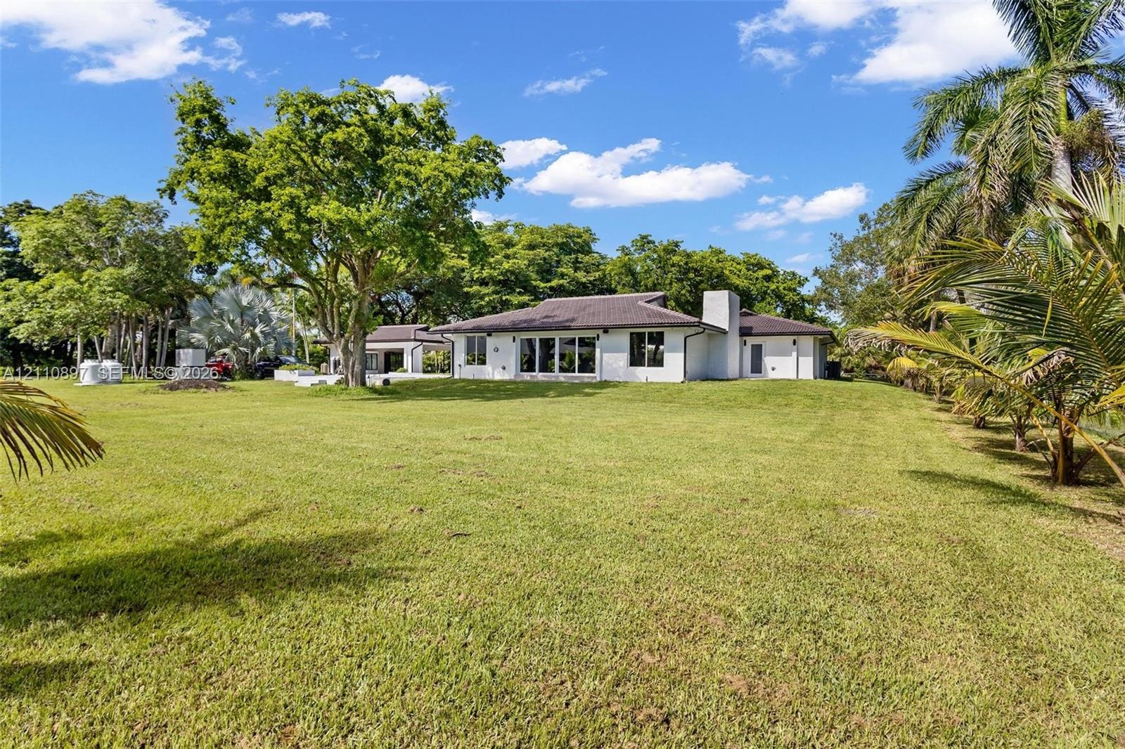 Single-story residence featuring a hipped tile roof, white stucco exterior, expansive lawn, mature specimen trees, and palm landscaping