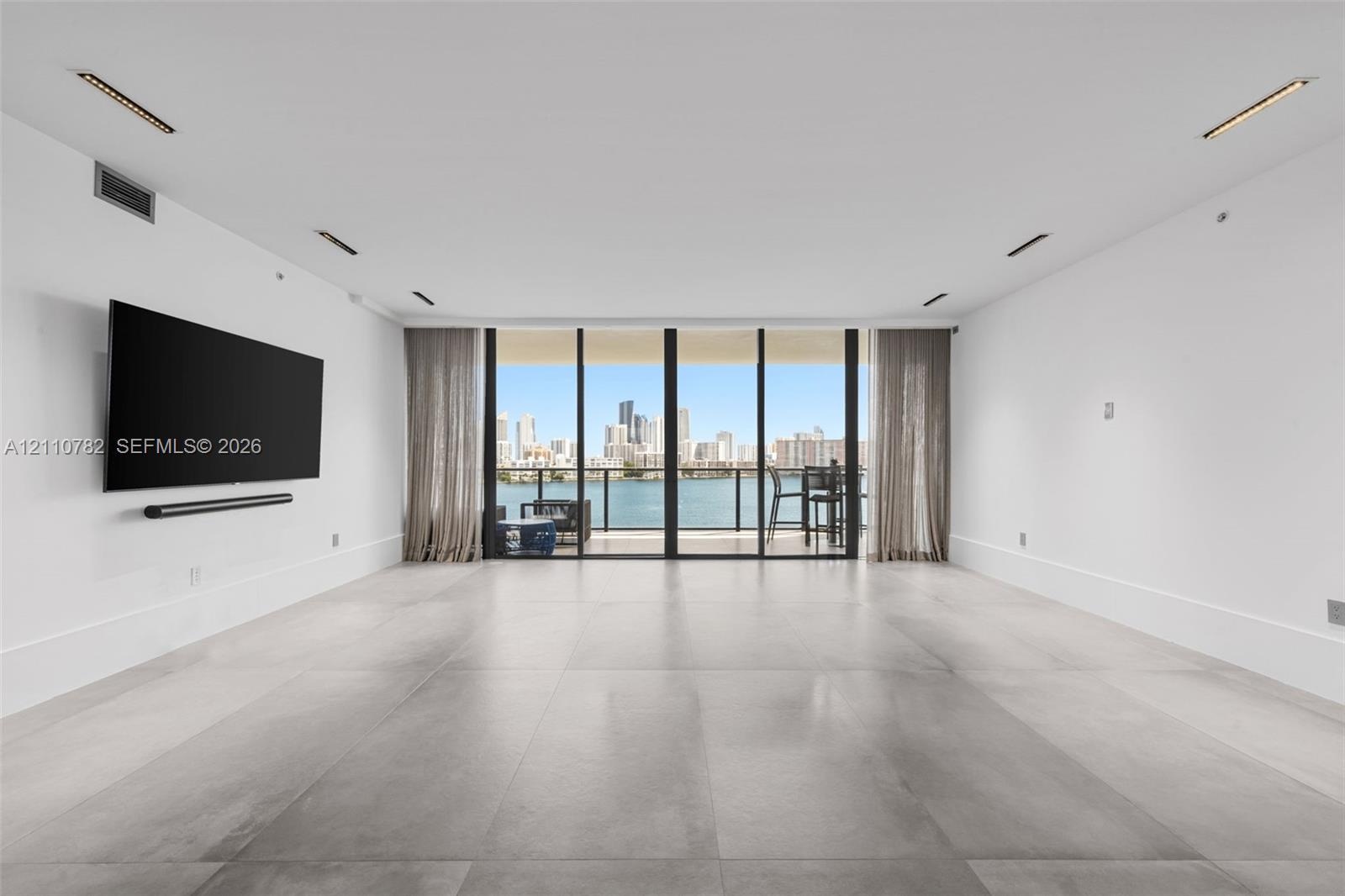 Large-format tile flooring, recessed linear lighting, and expansive floor-to-ceiling glass paneling leading to a balcony with water and skyline scener