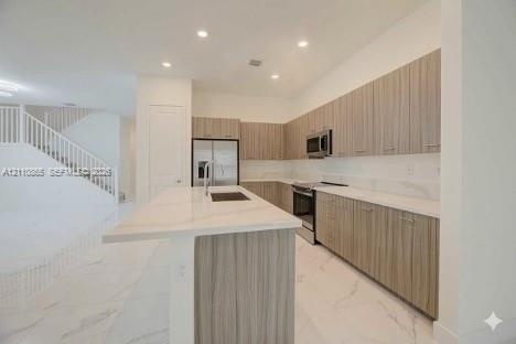 Kitchen featuring flat-panel wood-finish cabinetry, stone-surface countertops, a center island with an integrated sink, stainless steel appliances, an