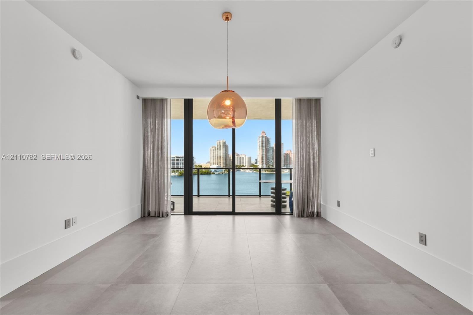Large-format tile flooring, oversized sliding glass door, spherical glass pendant light, and balcony with water and city skyline backdrop
