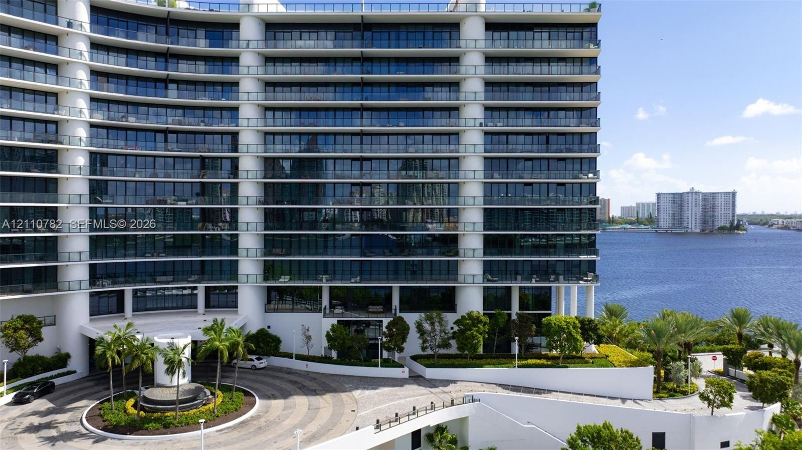 Mid-rise architectural exterior featuring tiered glass balconies, floor-to-ceiling windows, white structural columns, a circular paved motor court, an