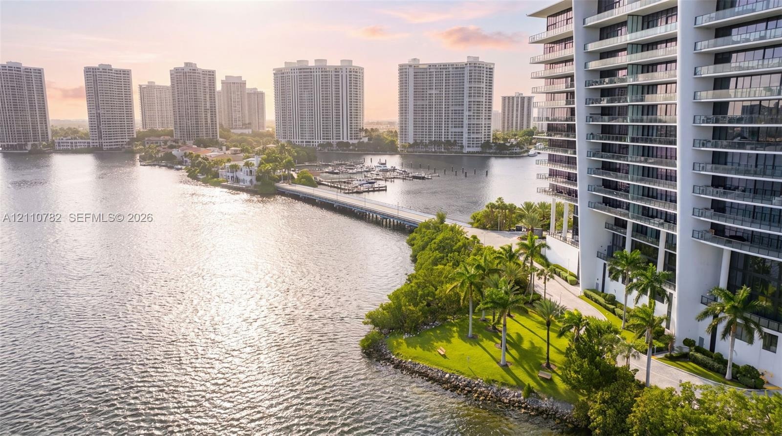 High-rise structure featuring stacked glass-railing balconies, situated on a waterfront lot with manicured lawn, mature palm trees, and direct bulkhea