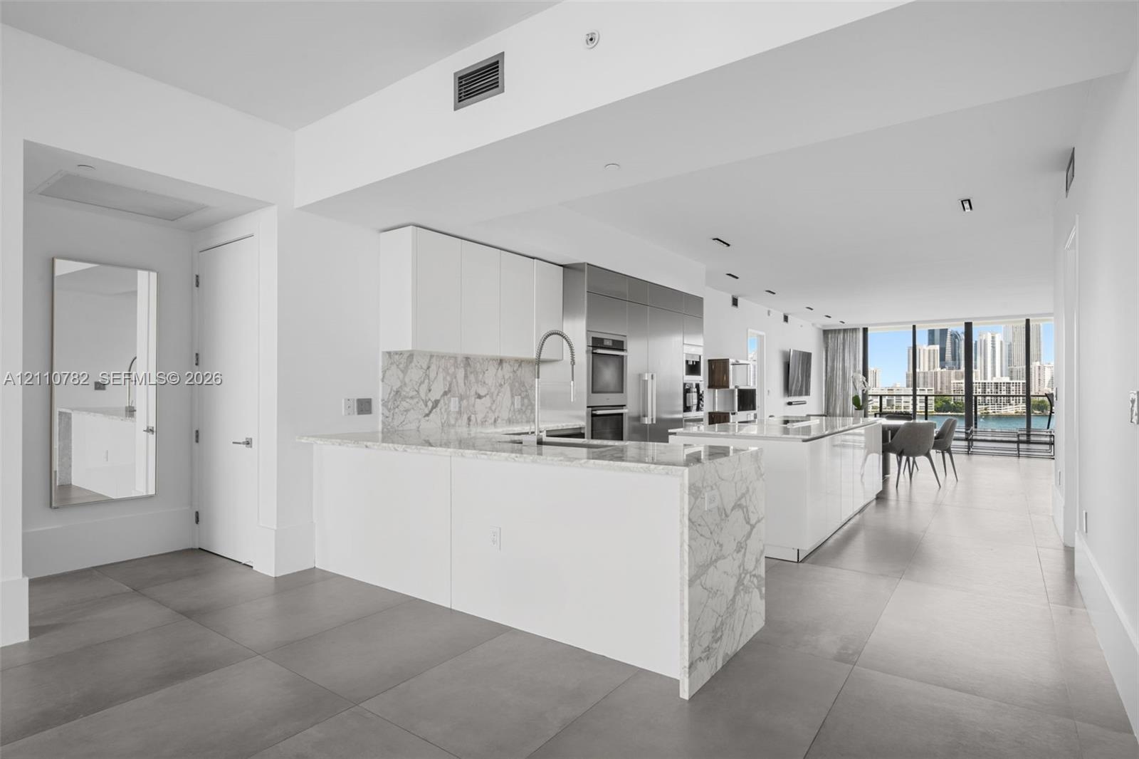 Kitchen featuring a stone-finish island with waterfall edge, white flat-panel cabinetry, stainless steel built-in appliances, and large-format floor t