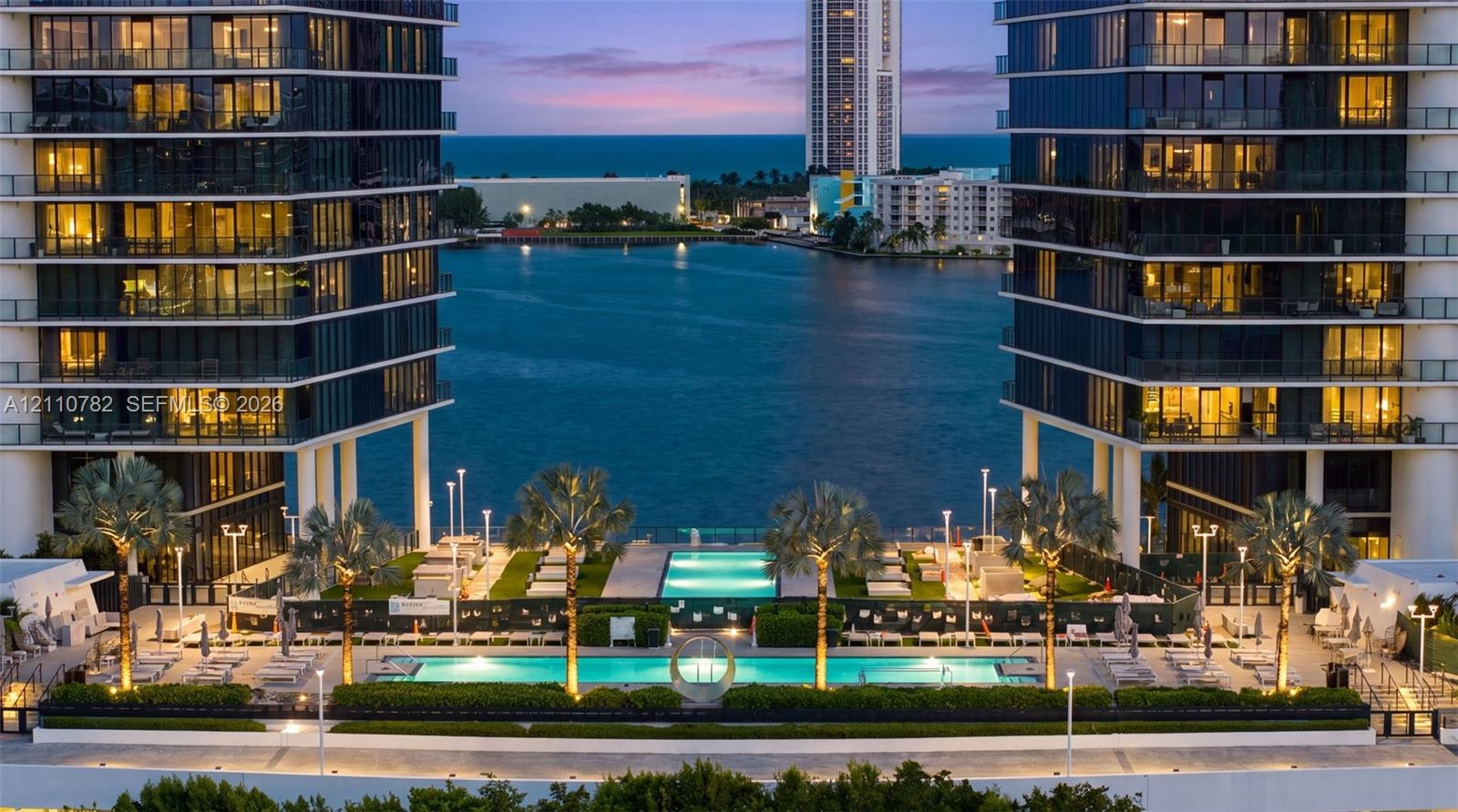 Waterfront high-rise complex featuring dual glass-facade towers, multiple swimming pools, professional landscaping, and paved outdoor terrace space