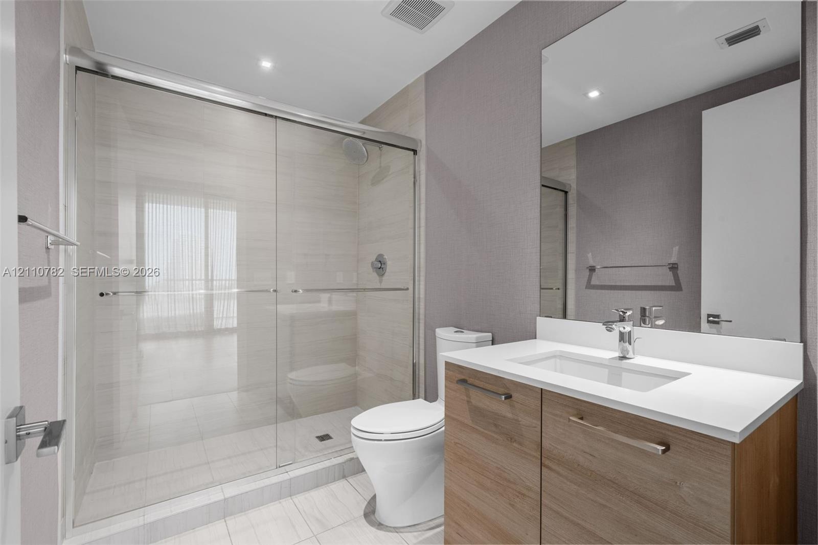 Bathroom featuring a glass-enclosed walk-in shower with tile surround, vanity with a light-tone wood finish, solid-surface countertop, and an integrat