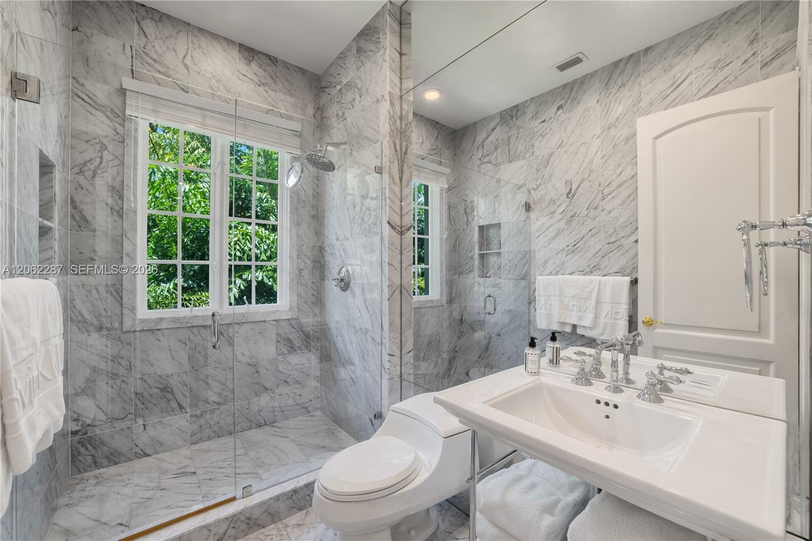 Full-wall natural stone tiling, glass-enclosed shower, wall-to-wall vanity mirror, porcelain sink with polished chrome widespread faucet, and multi-pa