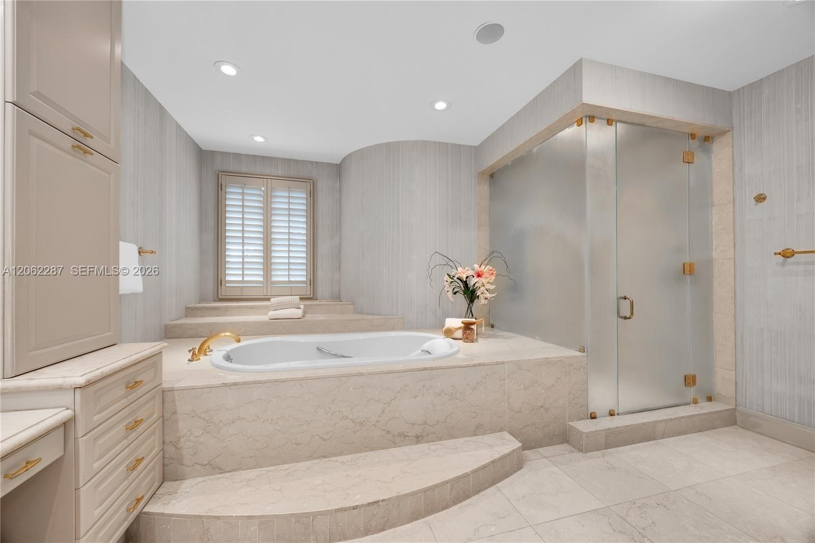 Raised stone-surround soaking tub with gold-tone fixtures, walk-in glass-enclosed shower, custom built-in cabinetry, floor-to-ceiling stone wall cladd