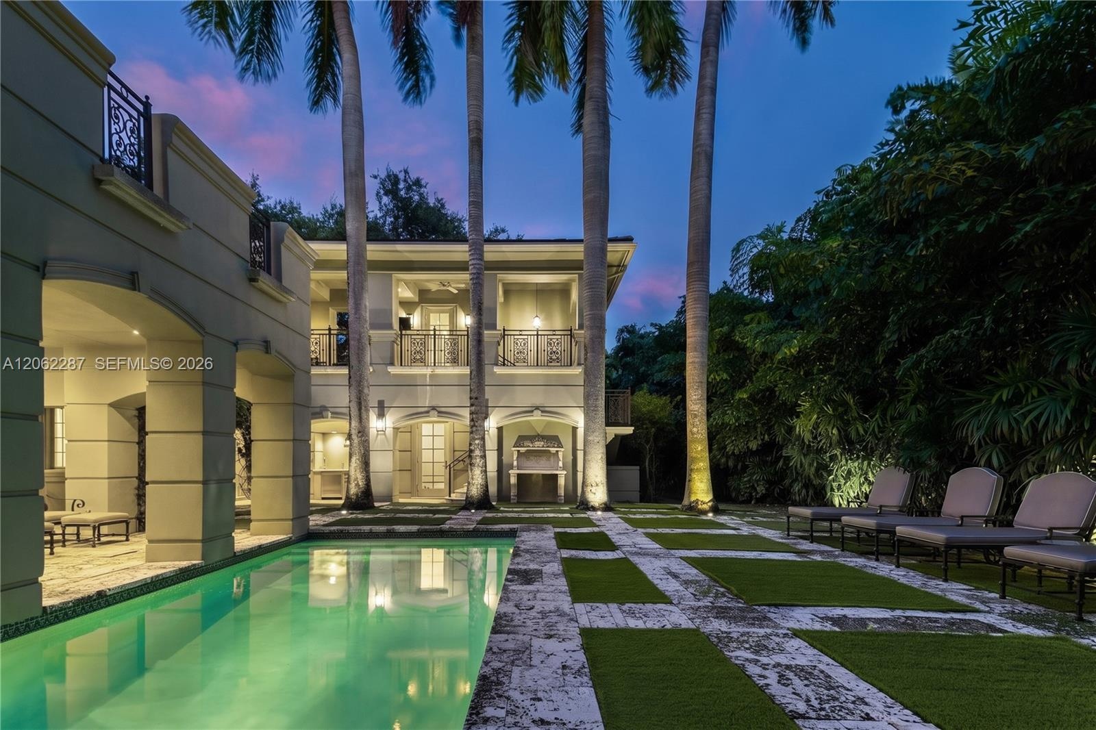 Exterior space featuring a rectangular swimming pool, stone-paver hardscaping, grid-patterned turf landscaping, mature palm trees, and two-story struc
