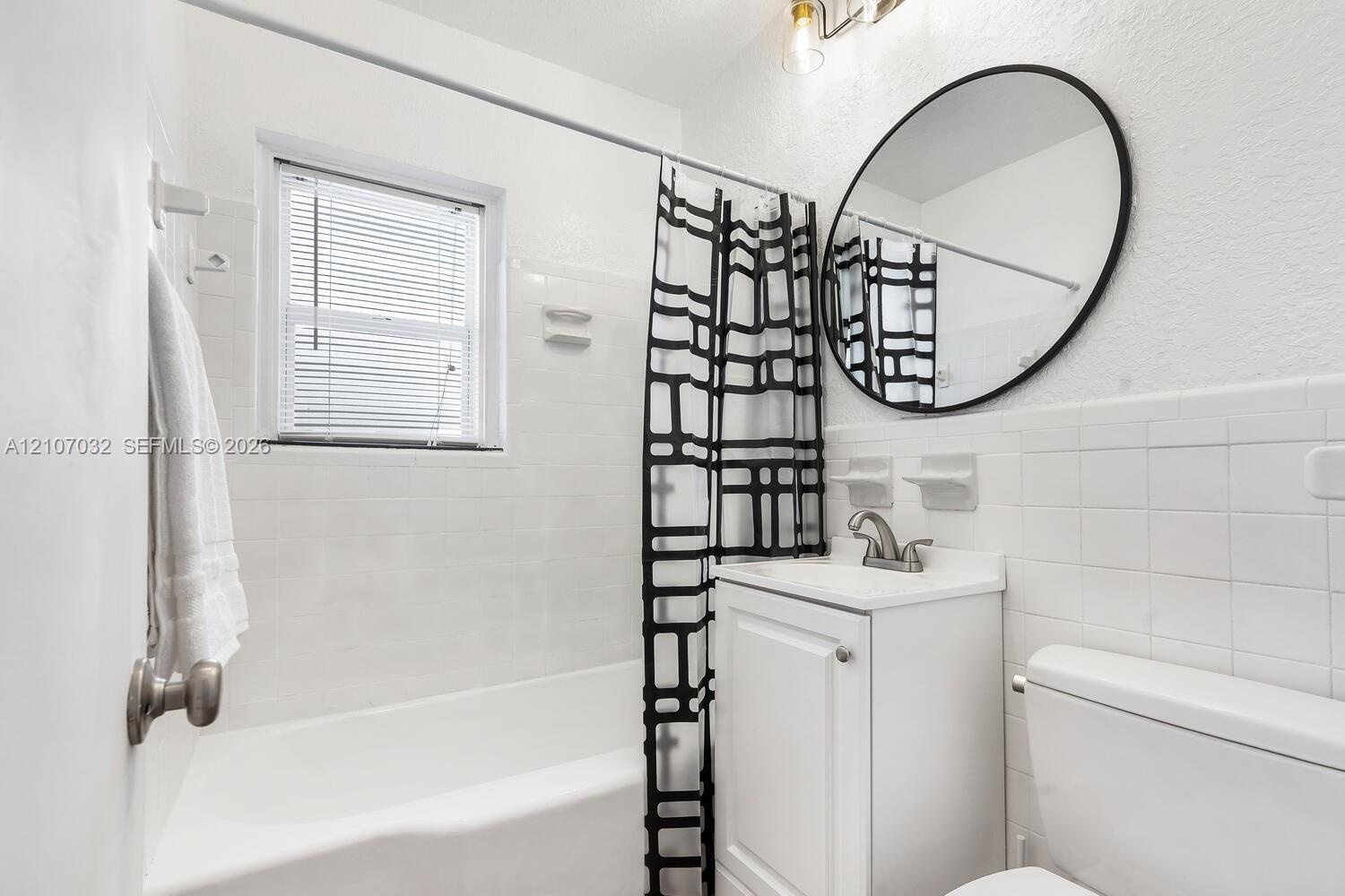 Bathroom featuring a white vanity cabinet, square wall tile surround, circular mirror with a black frame, and a tub-shower combination