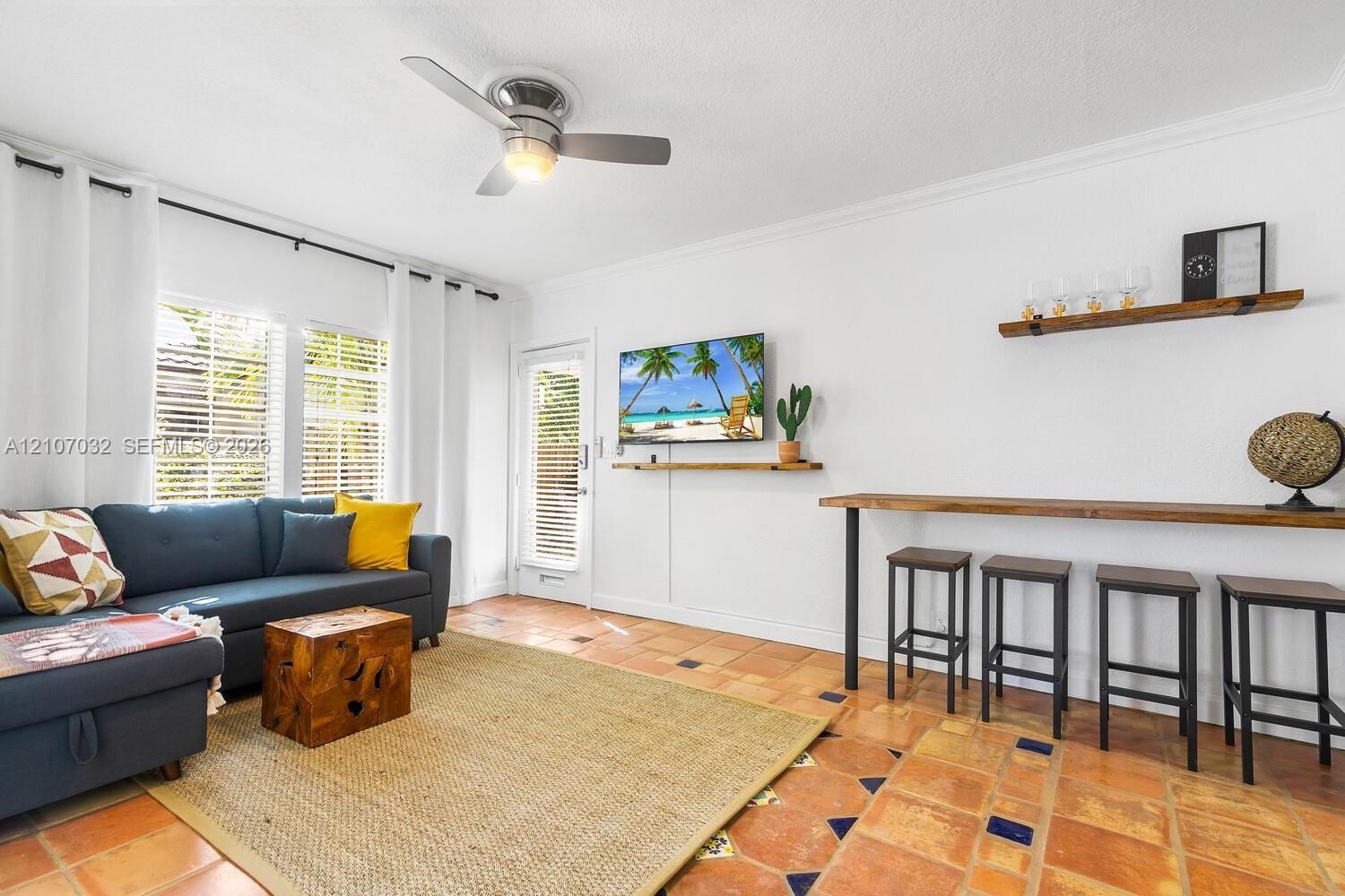 Living space featuring terracotta tile flooring with decorative tile inlays, a mounted ceiling fan, wall-mounted natural wood shelving, and a full-hei