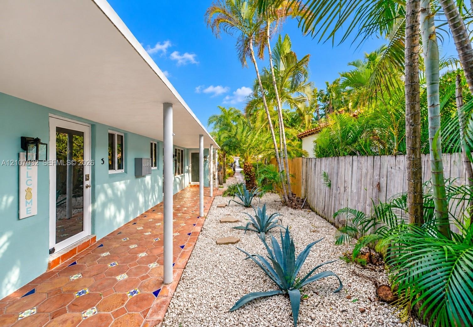 Covered walkway featuring terra cotta tile flooring with decorative inlay, light-blue exterior walls, support columns, and xeriscaped garden beds with