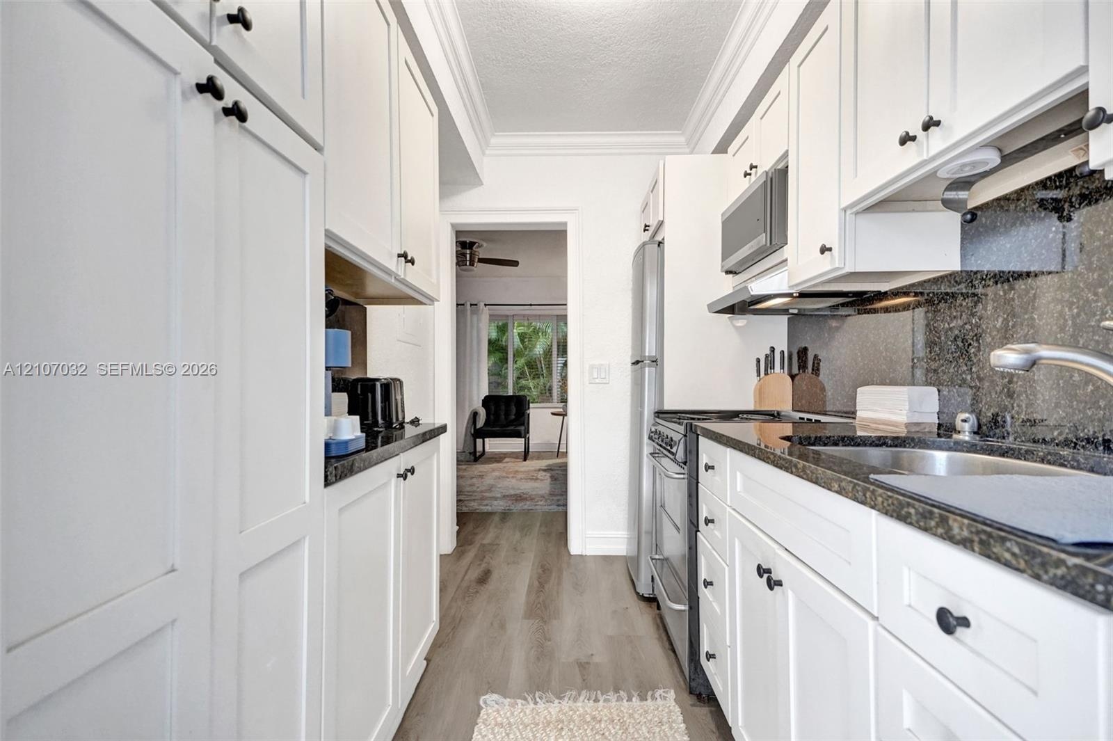 Galley kitchen featuring white shaker-style cabinetry, stone countertops, integrated stainless steel appliances, dark stone backsplash, and wood-finis