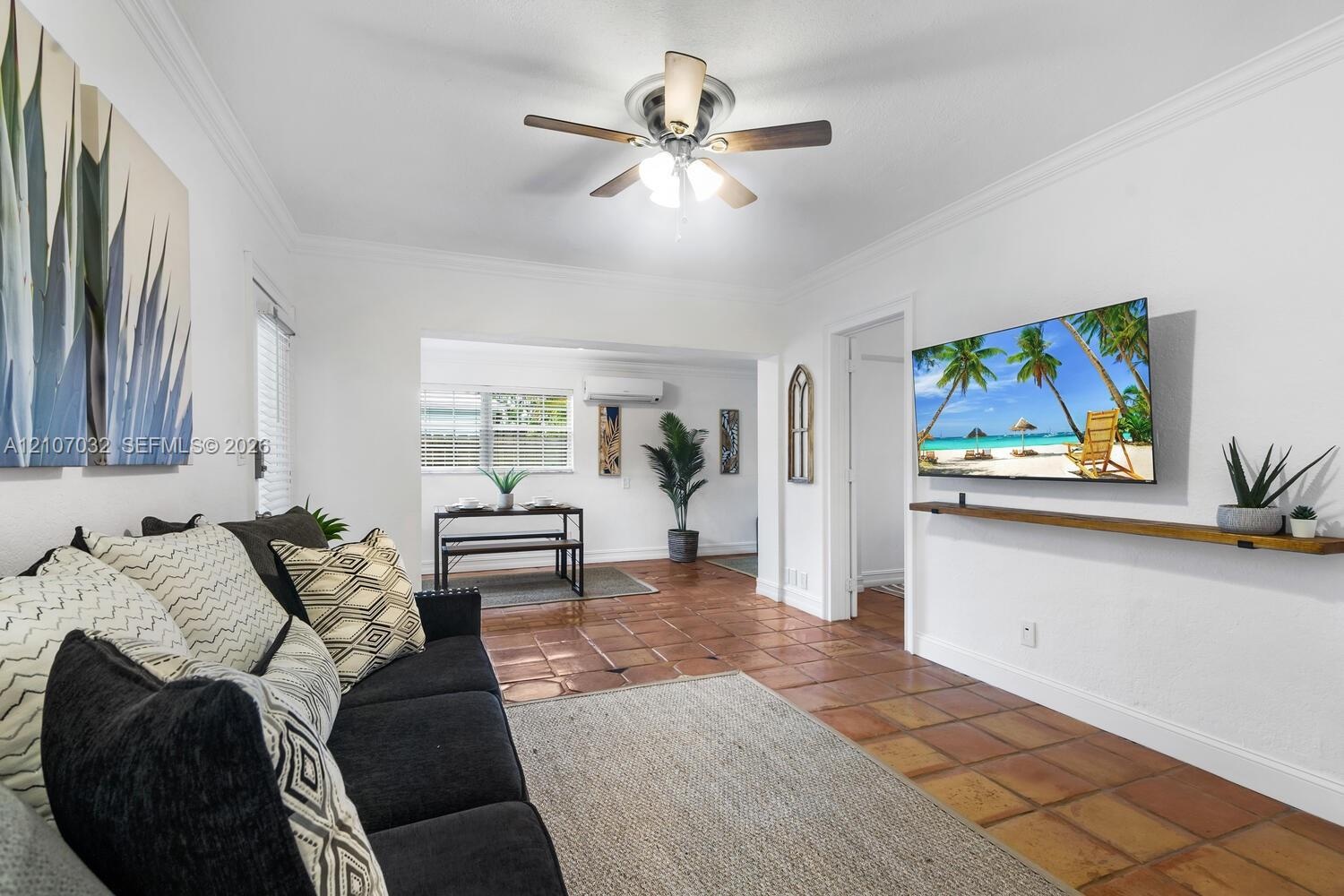 Living space featuring terracotta tile flooring, crown molding, a central ceiling fan with integrated lighting, and a wall-mounted wooden display shel