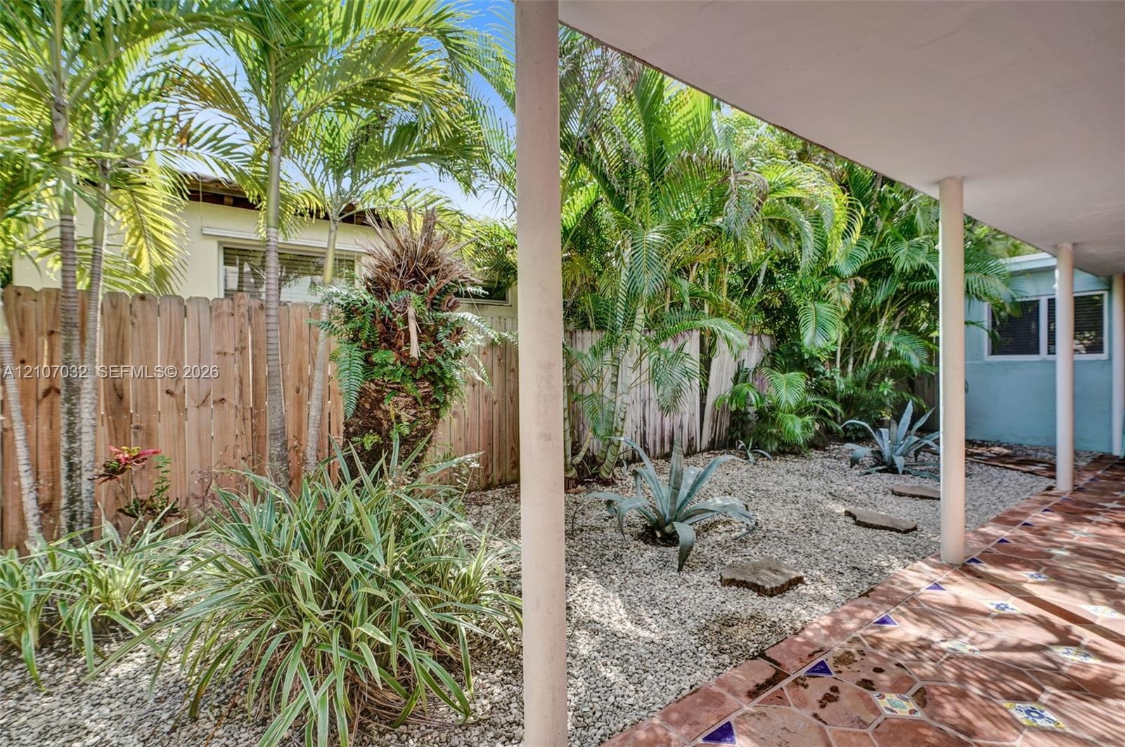 Covered patio featuring decorative tile flooring, gravel landscaping, established palms, and a wood privacy fence