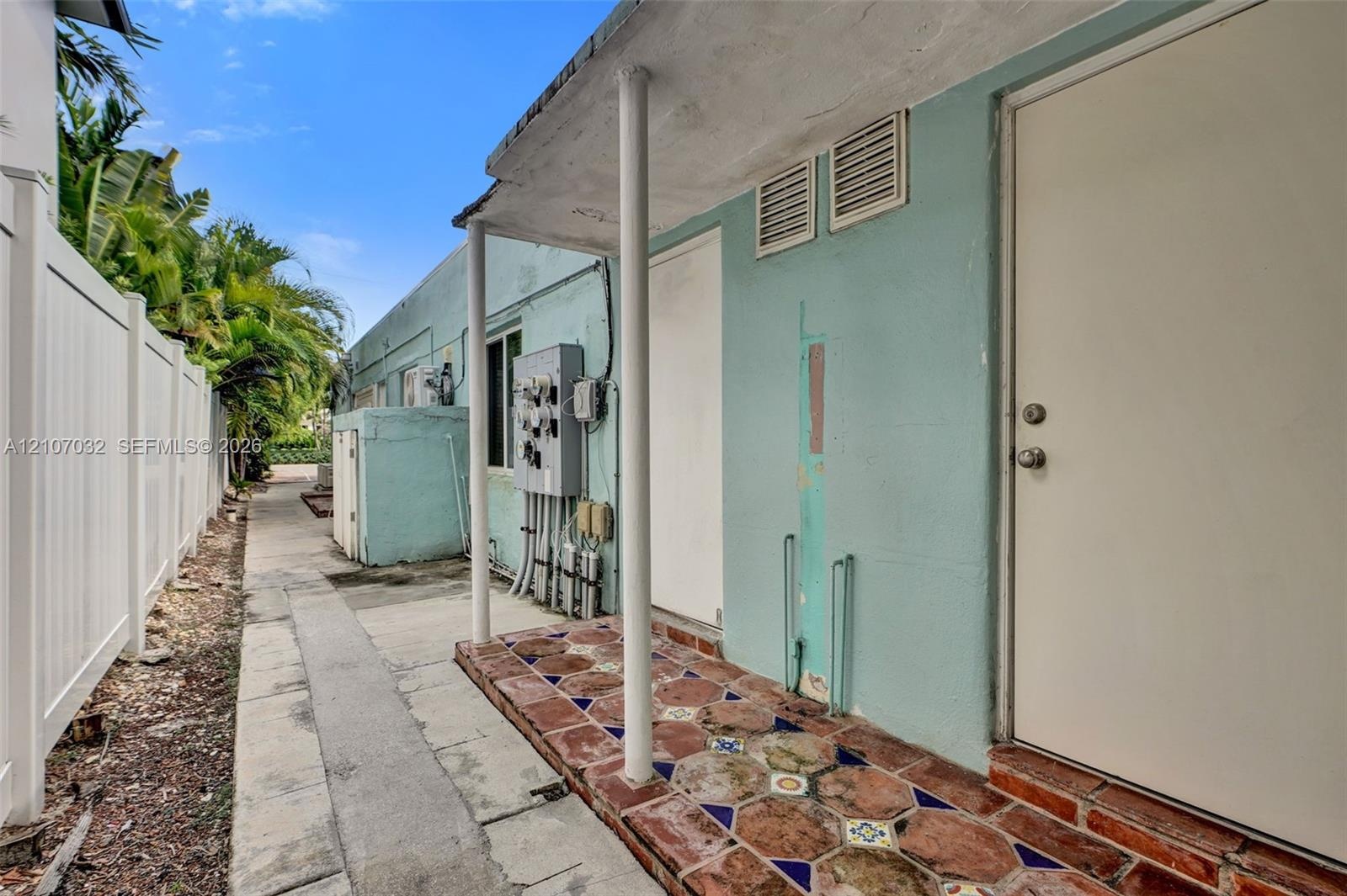 Exterior side entry featuring a covered porch with decorative patterned tile flooring, supported by round structural columns and enclosed by a tall pr
